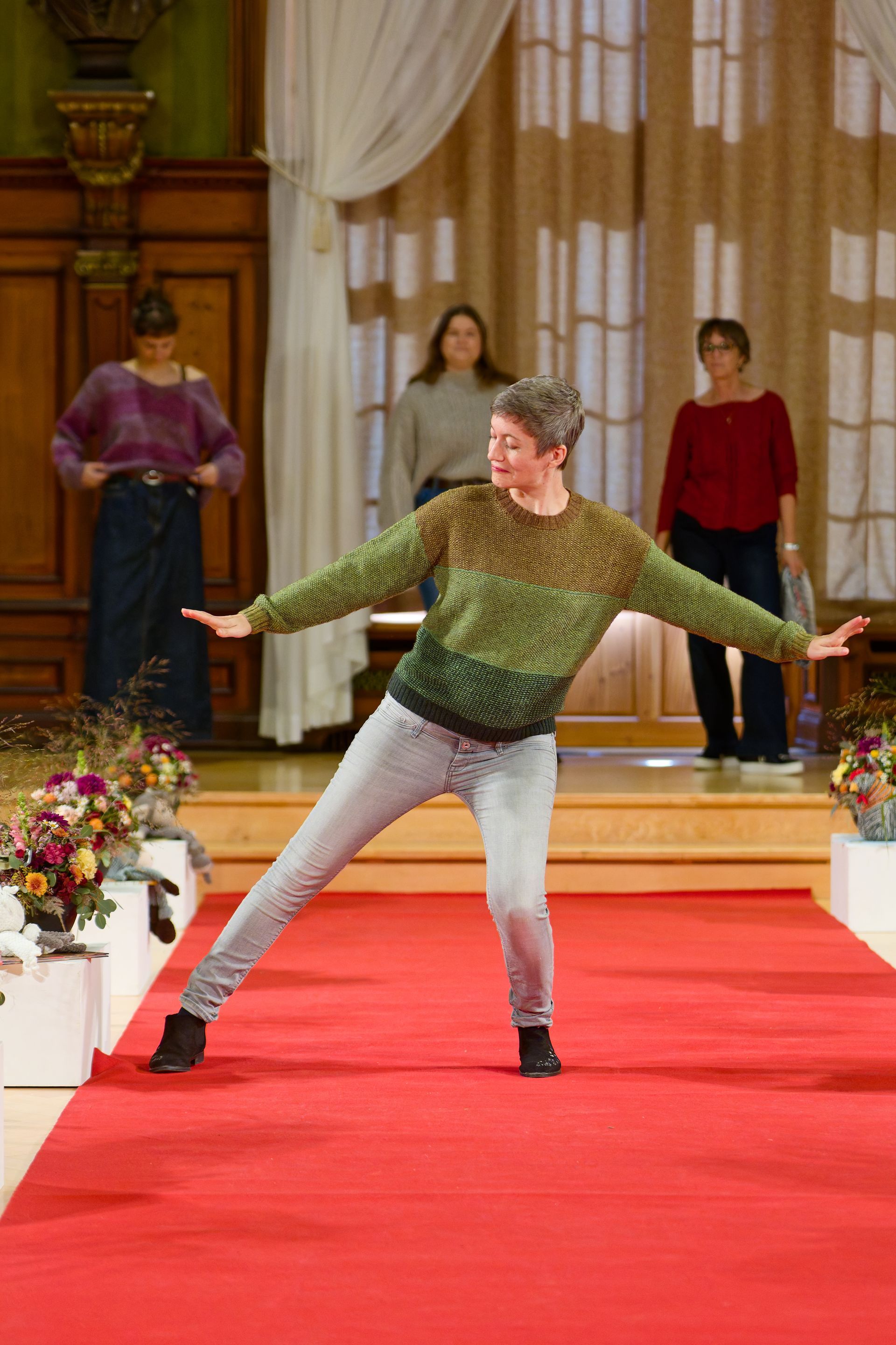 Person tanzt auf einem roten Teppich, sie trägt einen grünen Ombré-Pullover und Jeans, im Hintergrund stehen andere Personen.