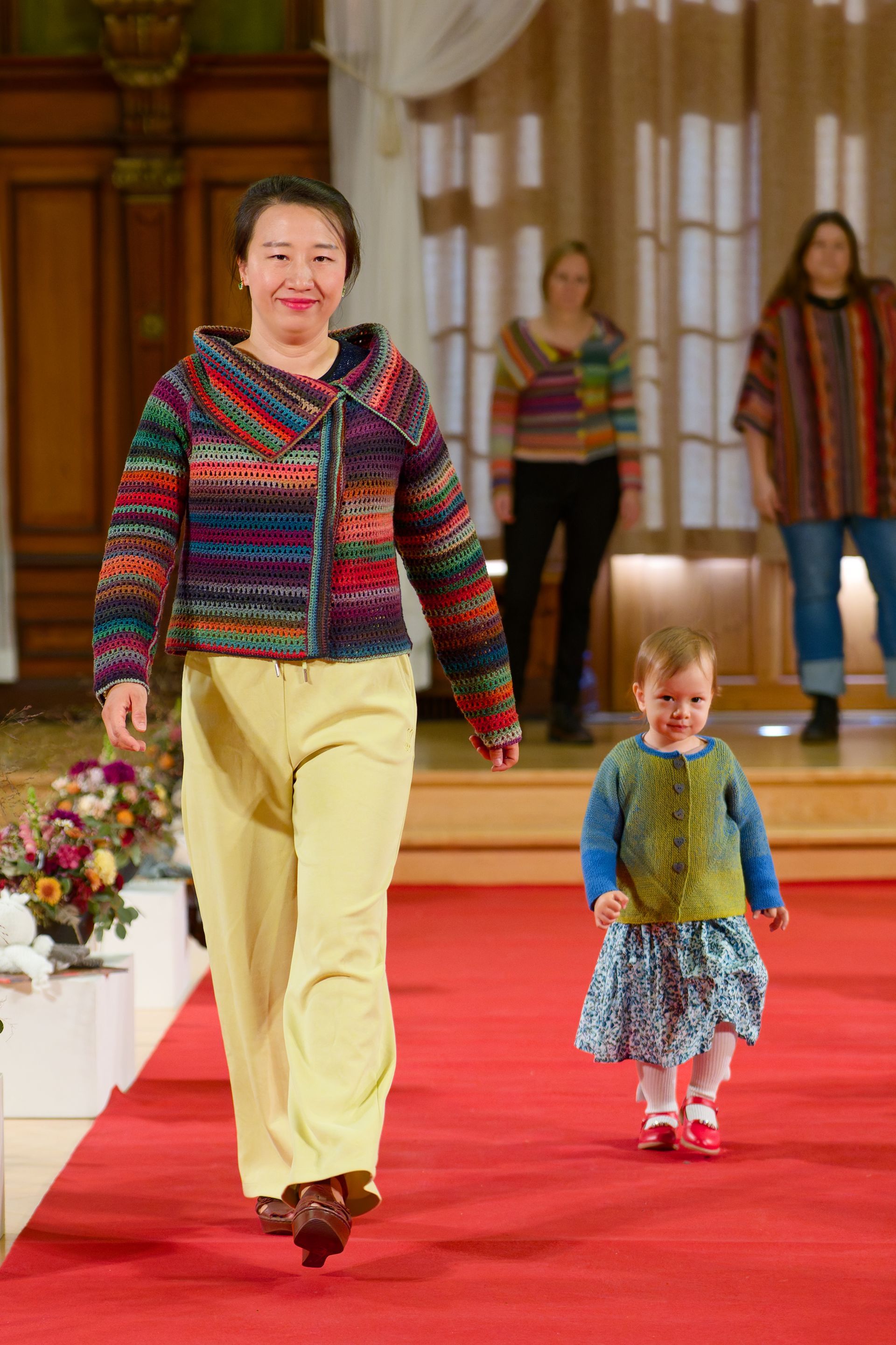Eine Frau in einer farbenfrohen Jacke und gelben Hosen läuft mit einem Kind in einem ähnlichen Pullover über den Laufsteg.