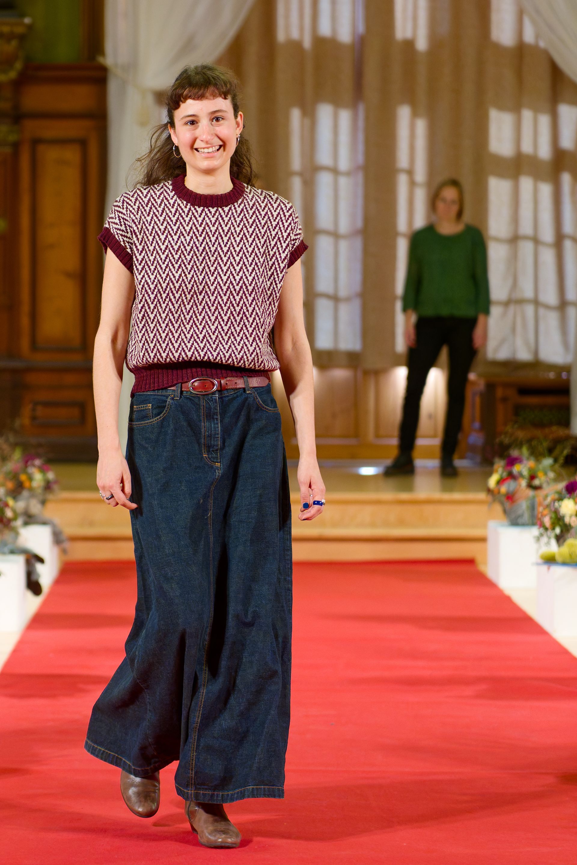 Eine Frau in einem gemusterten Oberteil und einem Jeansrock läuft über einen roten Laufsteg.