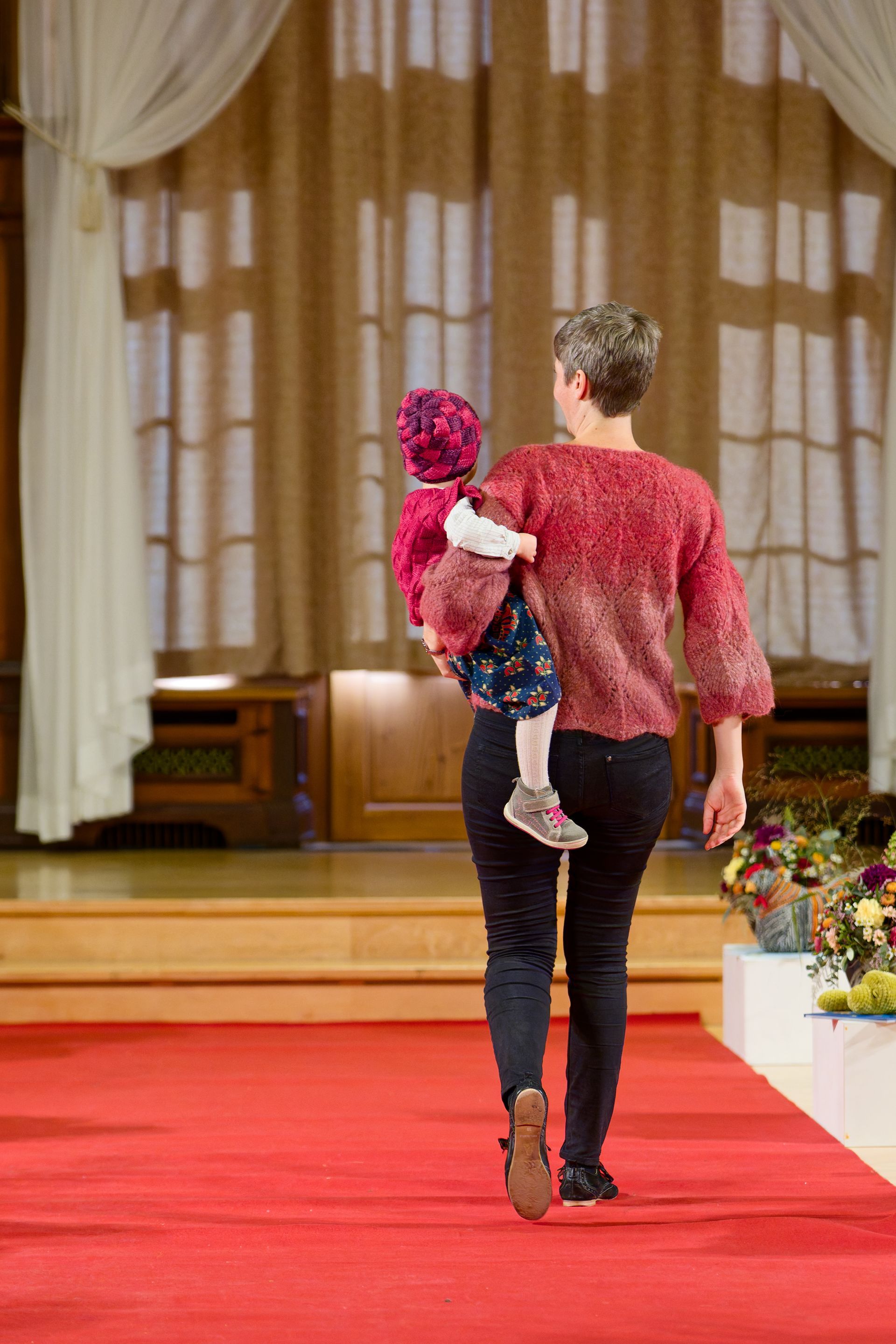 Eine Frau mit einem Kind auf dem Arm schreitet über einen roten Teppich. Beide tragen farbenfrohe Kleidung.
