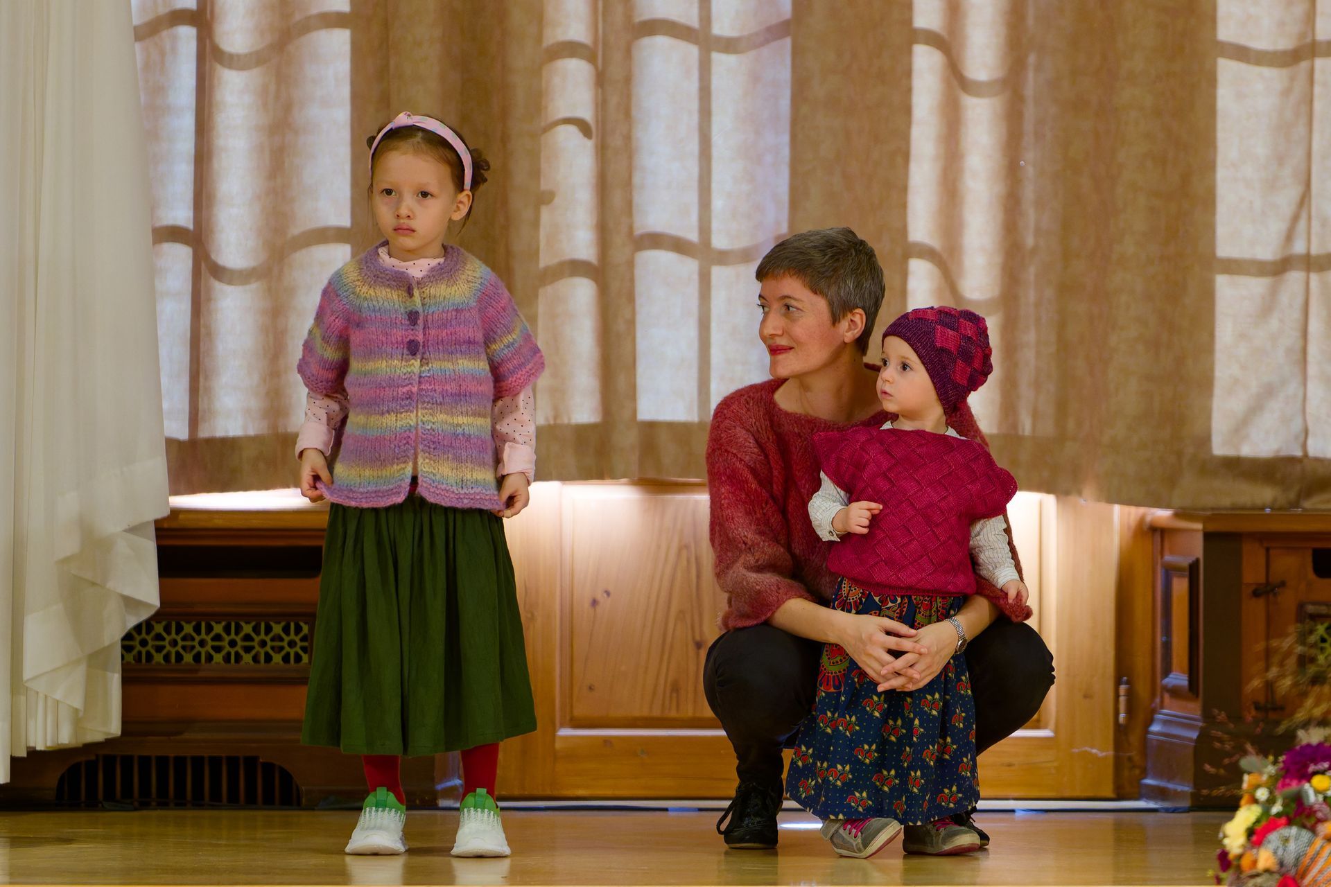 Eine Frau und zwei Kinder in Strickkleidung stehen vor einem Vorhang; eines der Kinder wird von der Frau gehalten.