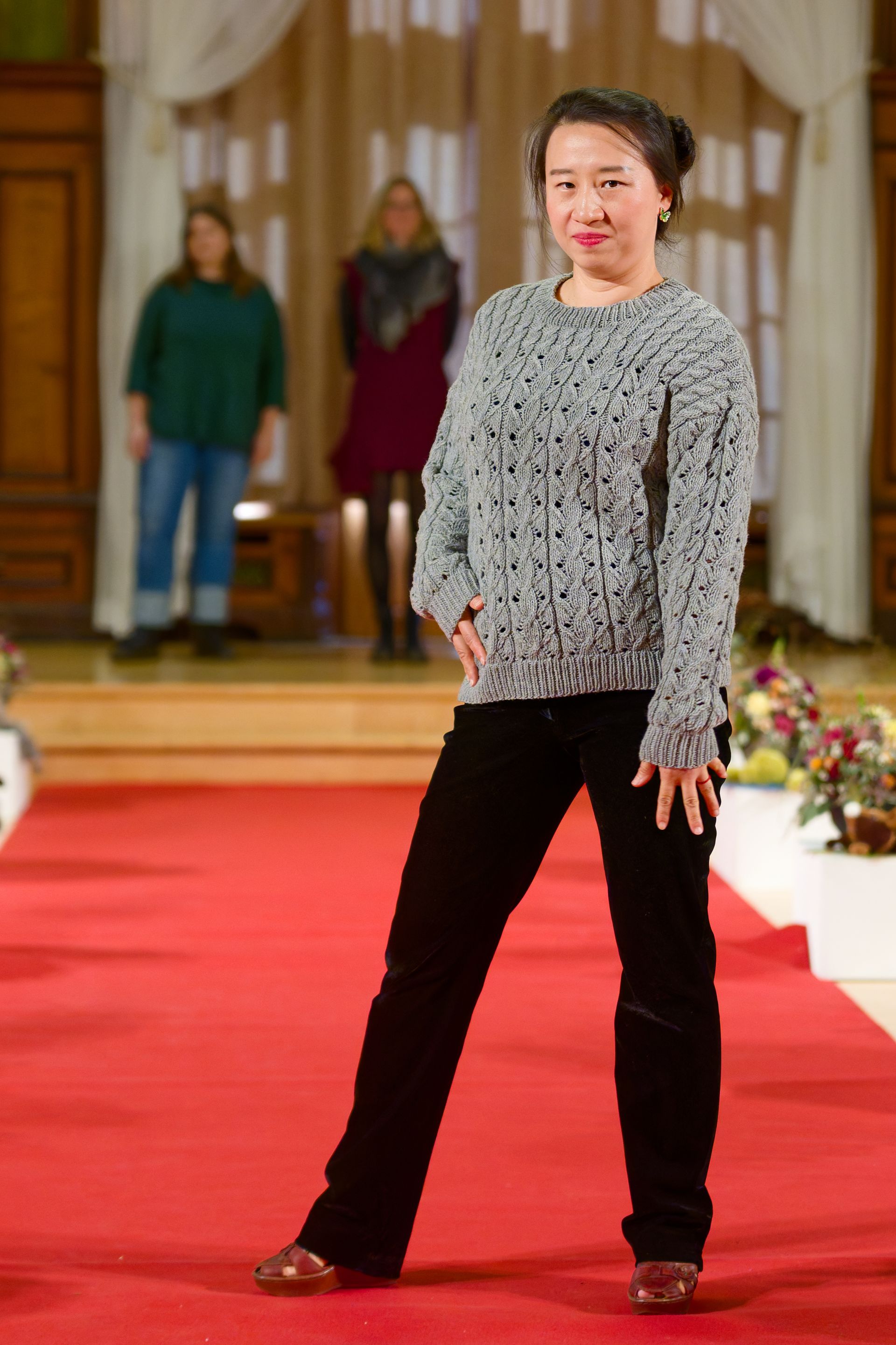 Frau in einem grauen Pullover und einer schwarzen Hose posiert auf einem roten Teppich; im Hintergrund stehen zwei Personen.
