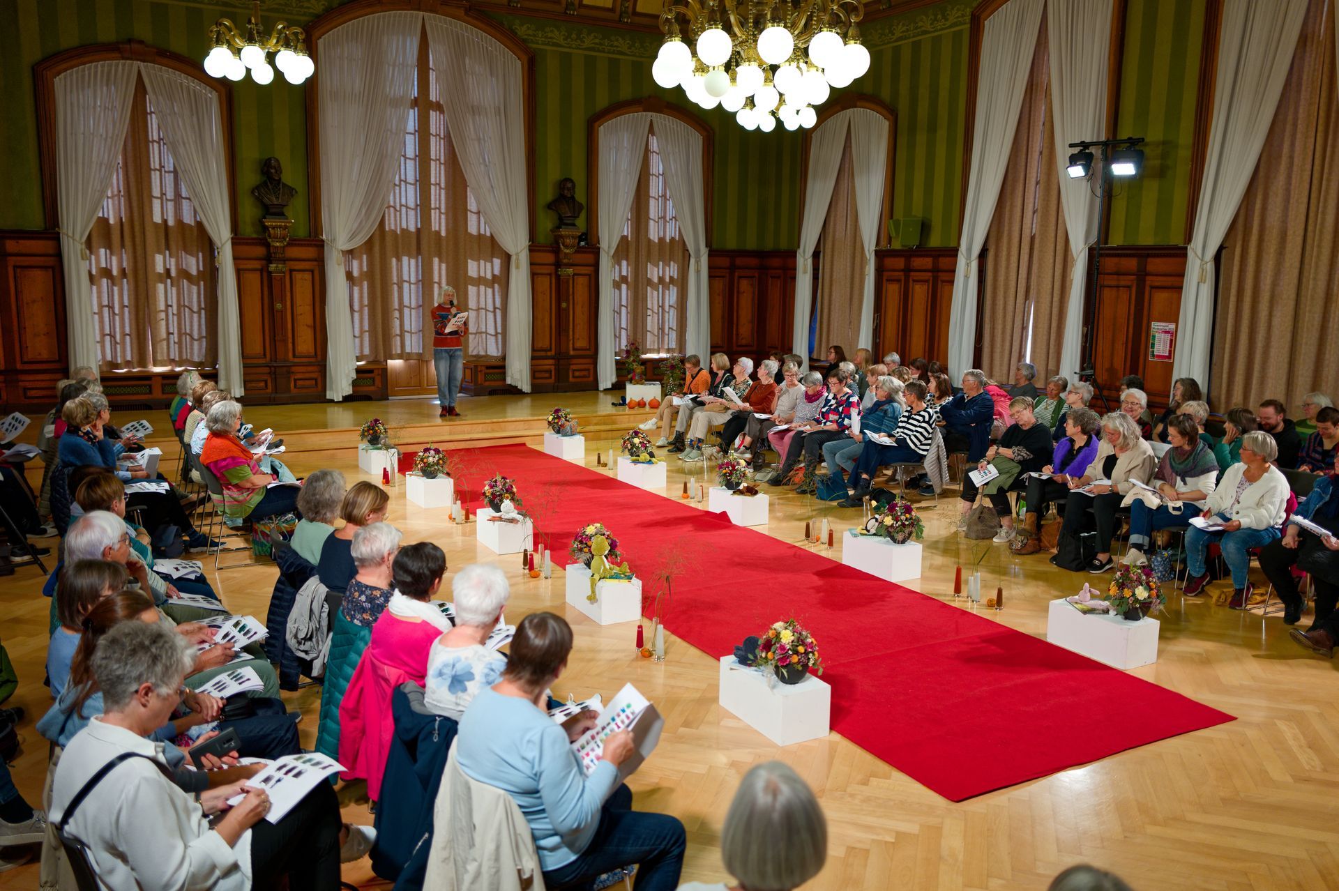 Das Publikum sitzt in einem großen Saal mit rotem Teppich und Ausstellungsstücken. Am Ende des Teppichs steht ein Redner.