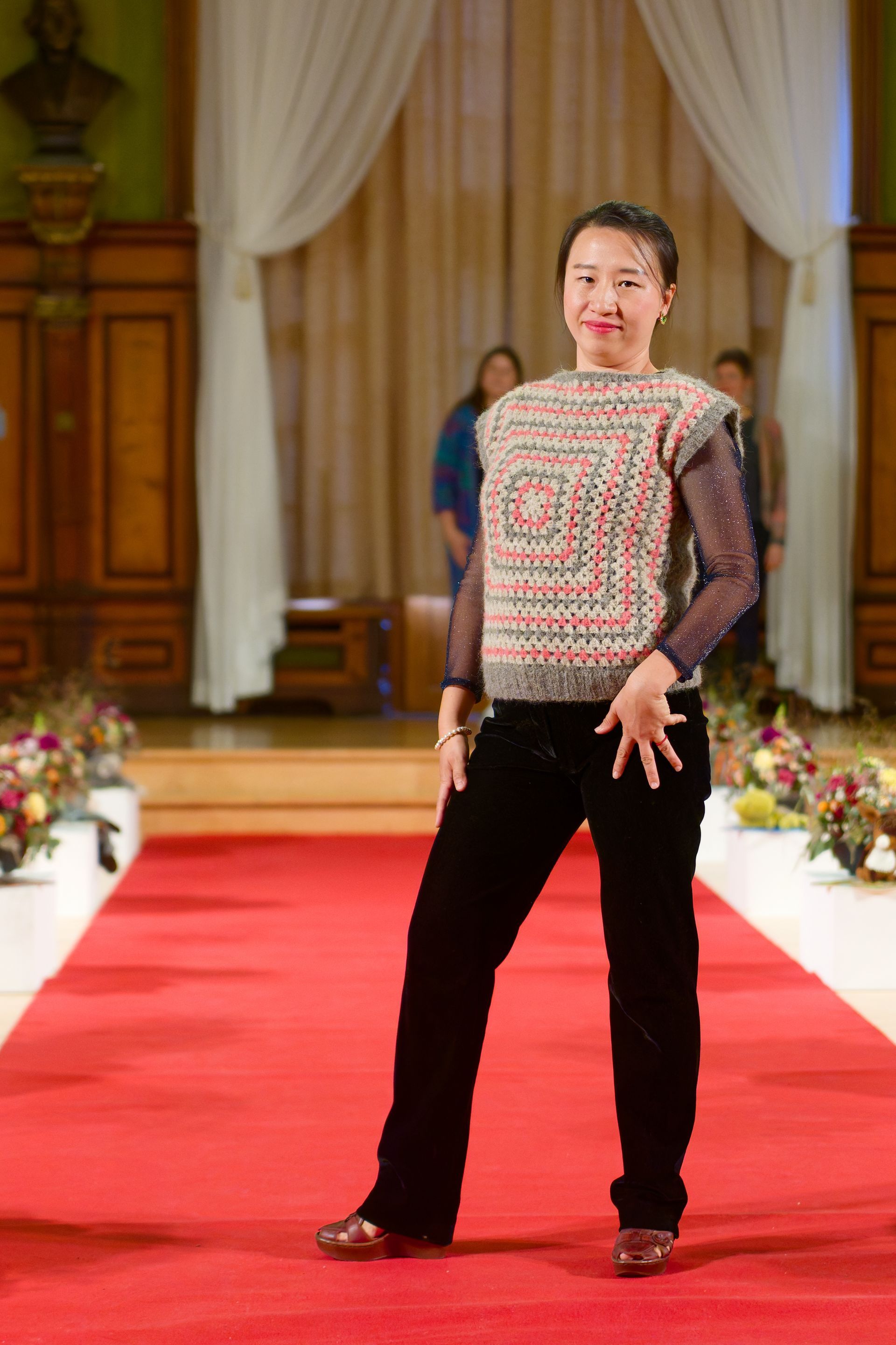 Frau in gemustertem Pullover und schwarzer Hose auf rotem Laufsteg.