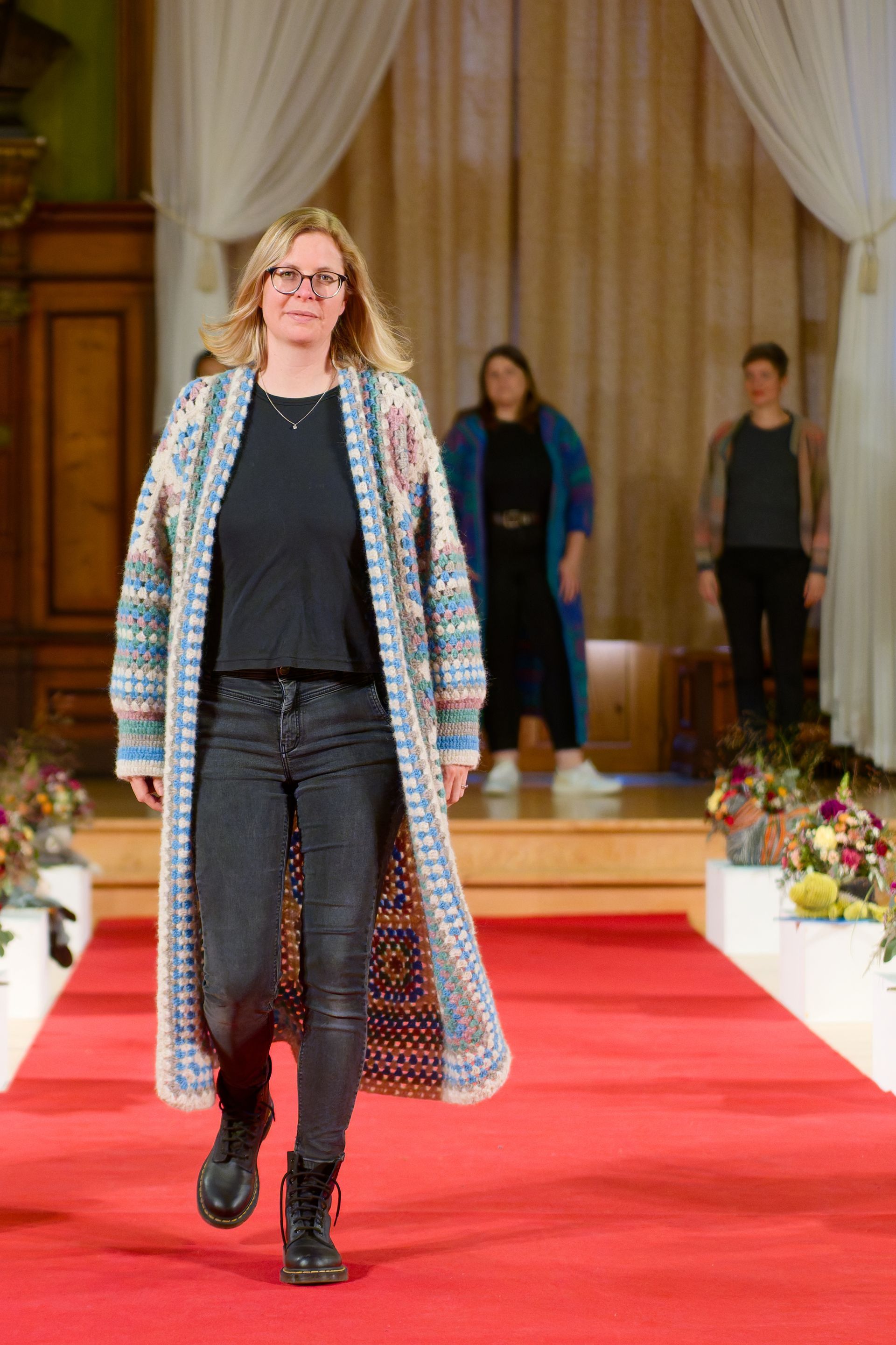 Eine Frau in einem langen, gemusterten Cardigan und schwarzen Stiefeln läuft über den Laufsteg.