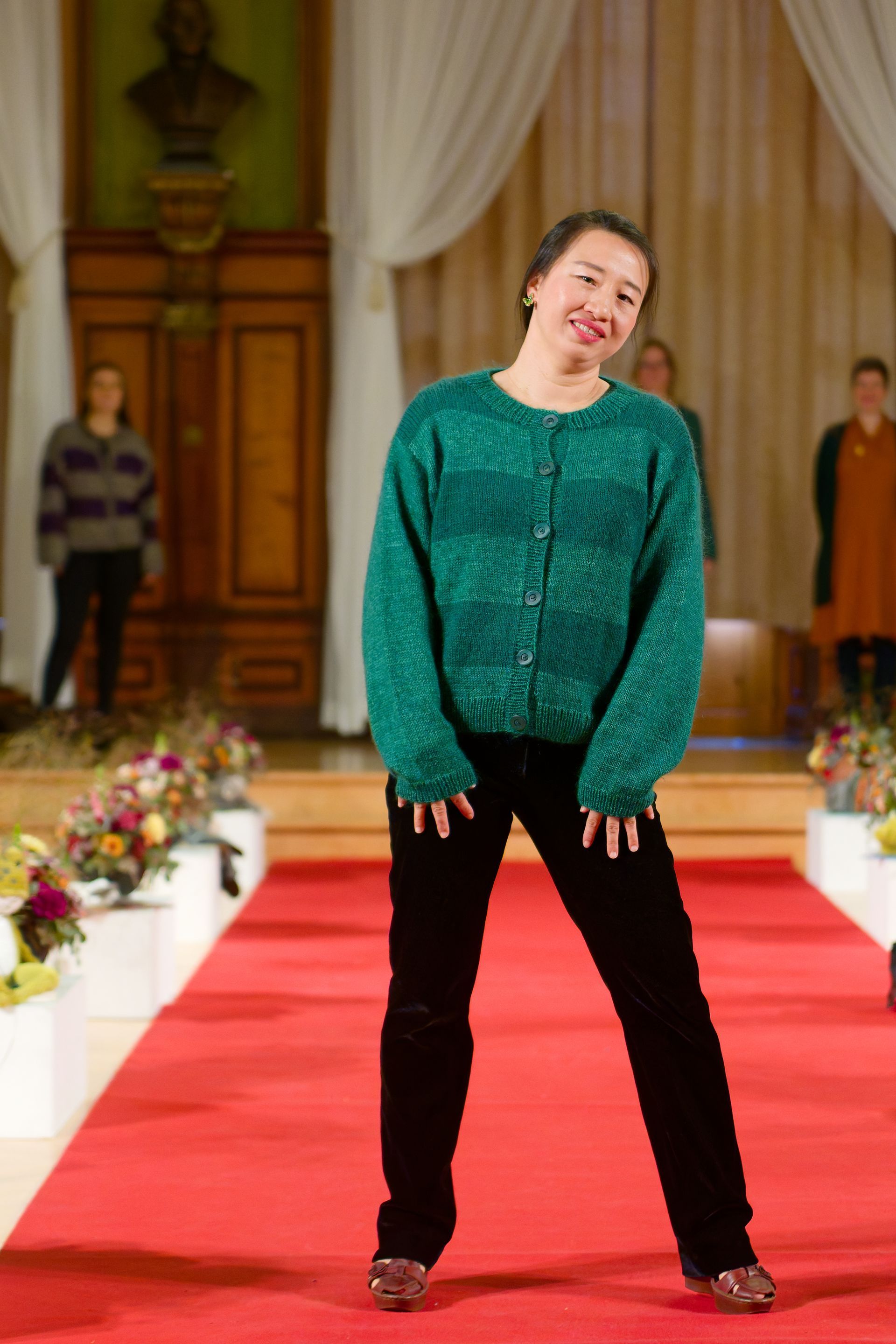 Frau auf dem Laufsteg in grünem Pullover und schwarzer Hose. Roter Teppich, Blumen, weitere Personen im Hintergrund.