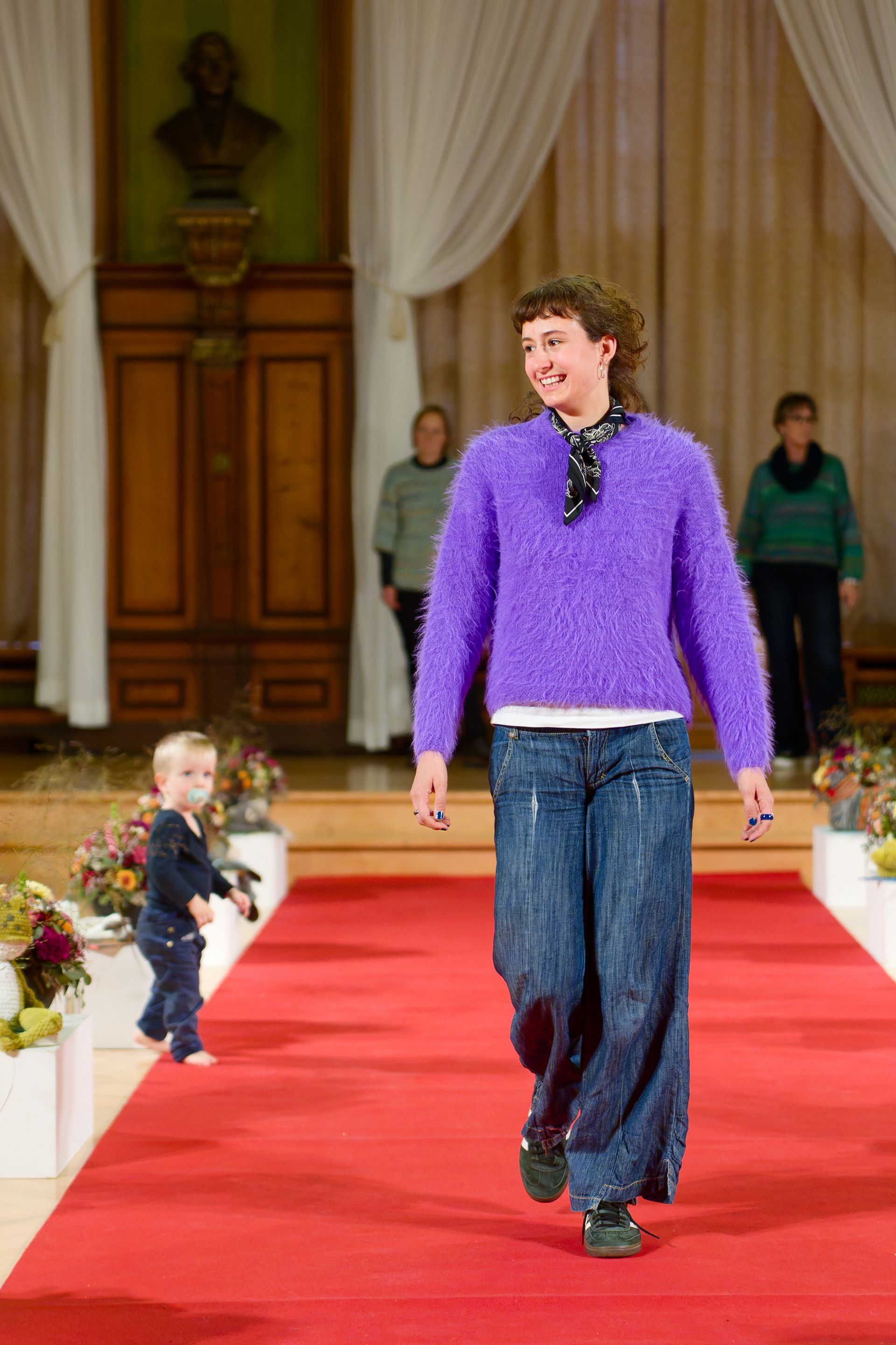 Person in lila Pullover + Jeans schreitet über den roten Teppich. Im Hintergrund sind ein Kind & 2 weitere Personen zu sehen.