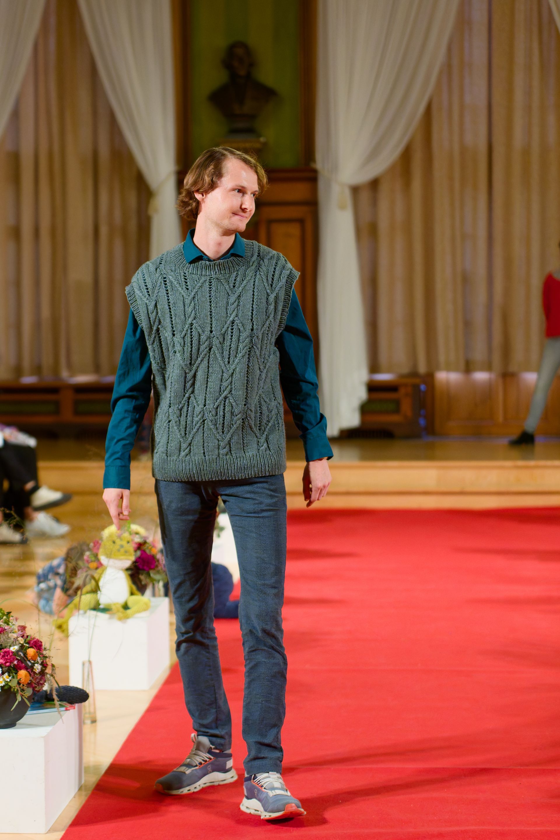 Ein Mann in türkisfarbener Strickweste, blauem Hemd und Jeans geht mit einem Spielzeug in der Hand über einen roten Teppich.