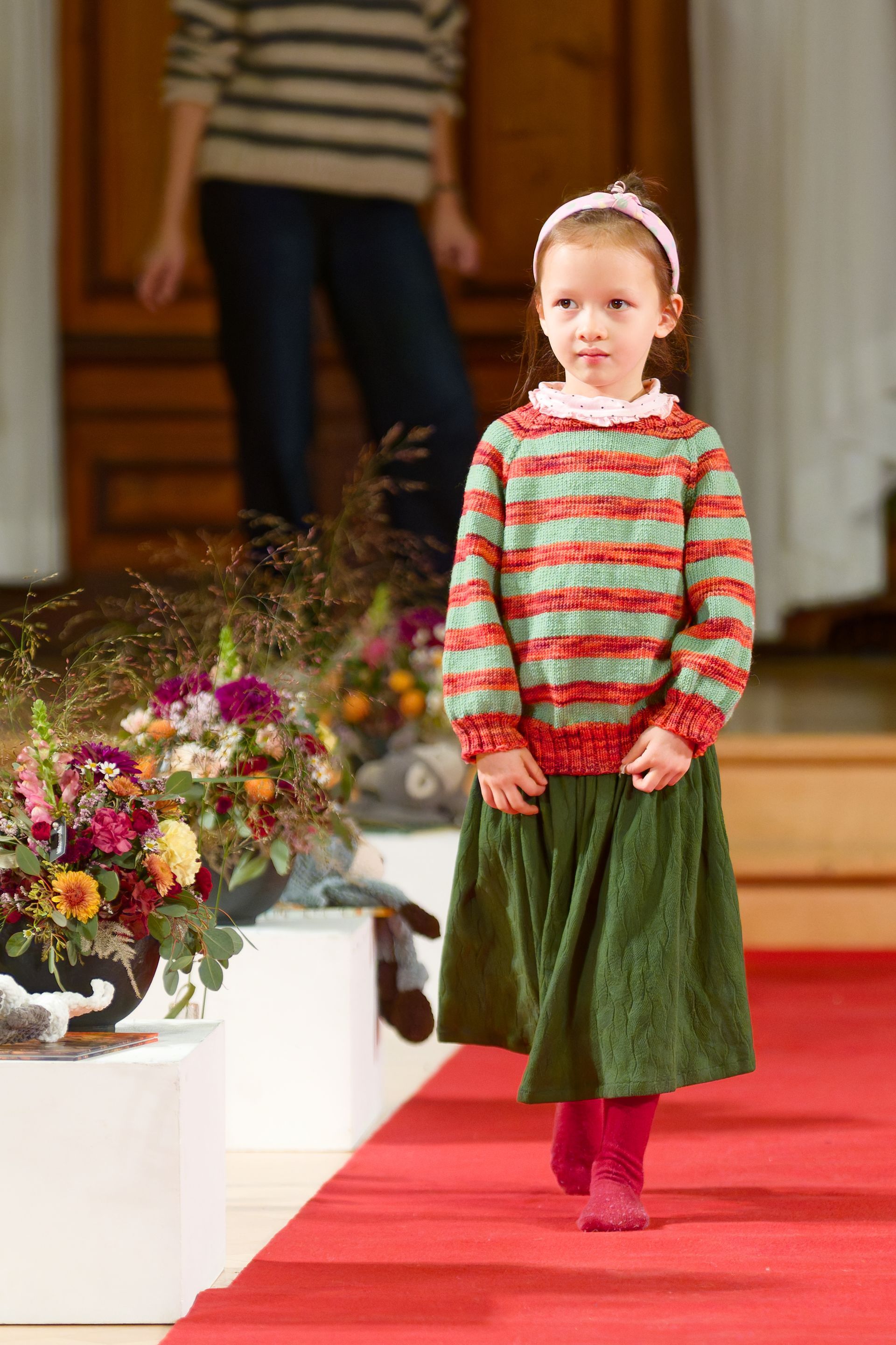Ein junges Kind in einem bunten Pullover und einem grünen Rock schreitet über einen roten Teppich.