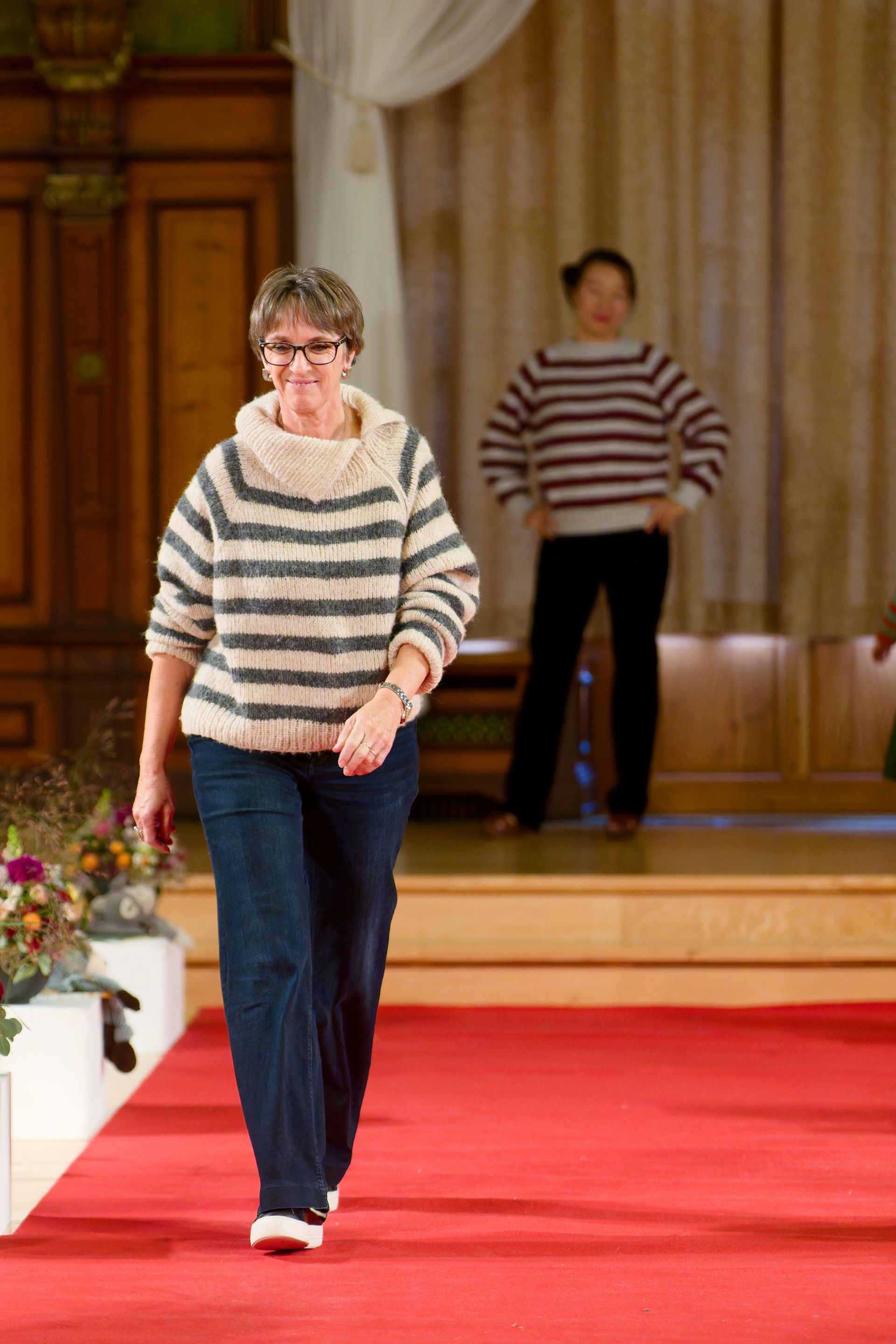 Frau in einem gestreiften Pullover schreitet über einen roten Teppich, eine andere Person steht hinter ihr auf einer Bühne.