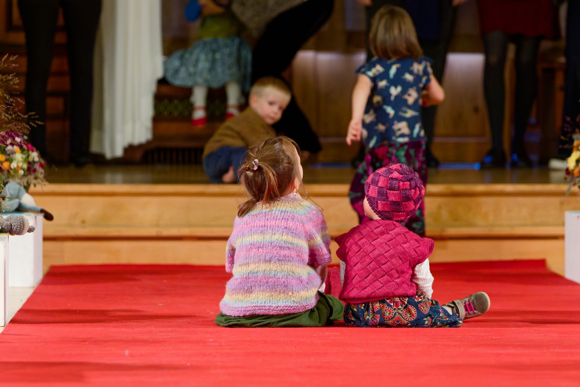 Kinder sitzen auf einem roten Teppich und blicken in Richtung Bühne. Im Hintergrund stehen Menschen.