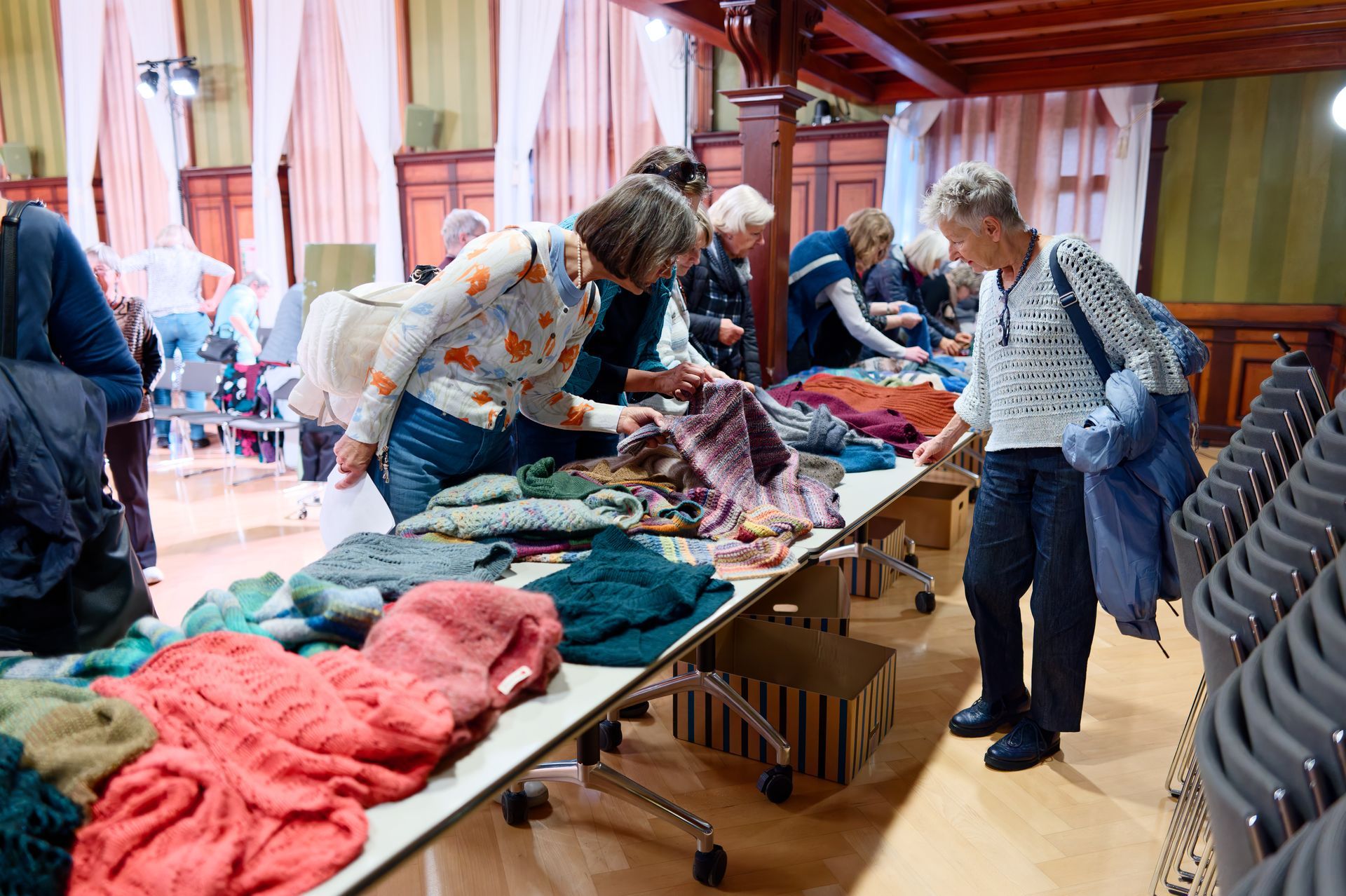 Menschen, die auf einem Kunsthandwerkermarkt in einer Halle an einem Tisch mit farbenfrohen Strickwaren stöbern.