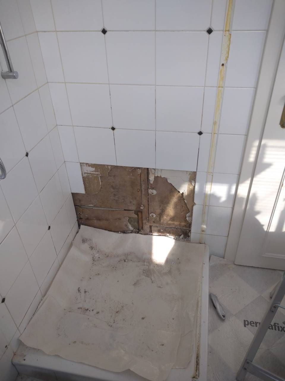 Mur de salle de bains dont des carreaux manquants laissent apparaître le mur brun en dessous. Les murs environnants sont carrelés en blanc.