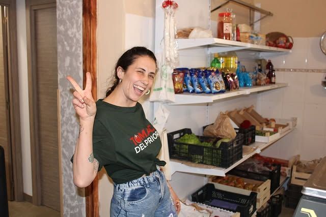 Mujer sonriendo, mostrando el símbolo de la paz frente a estantes repletos de aperitivos y productos frescos.