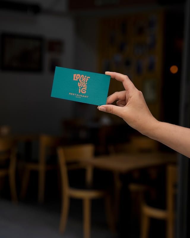 Mano sosteniendo una tarjeta color verde azulado con texto dorado dentro de un restaurante, fondo desenfocado con mesas y sillas.