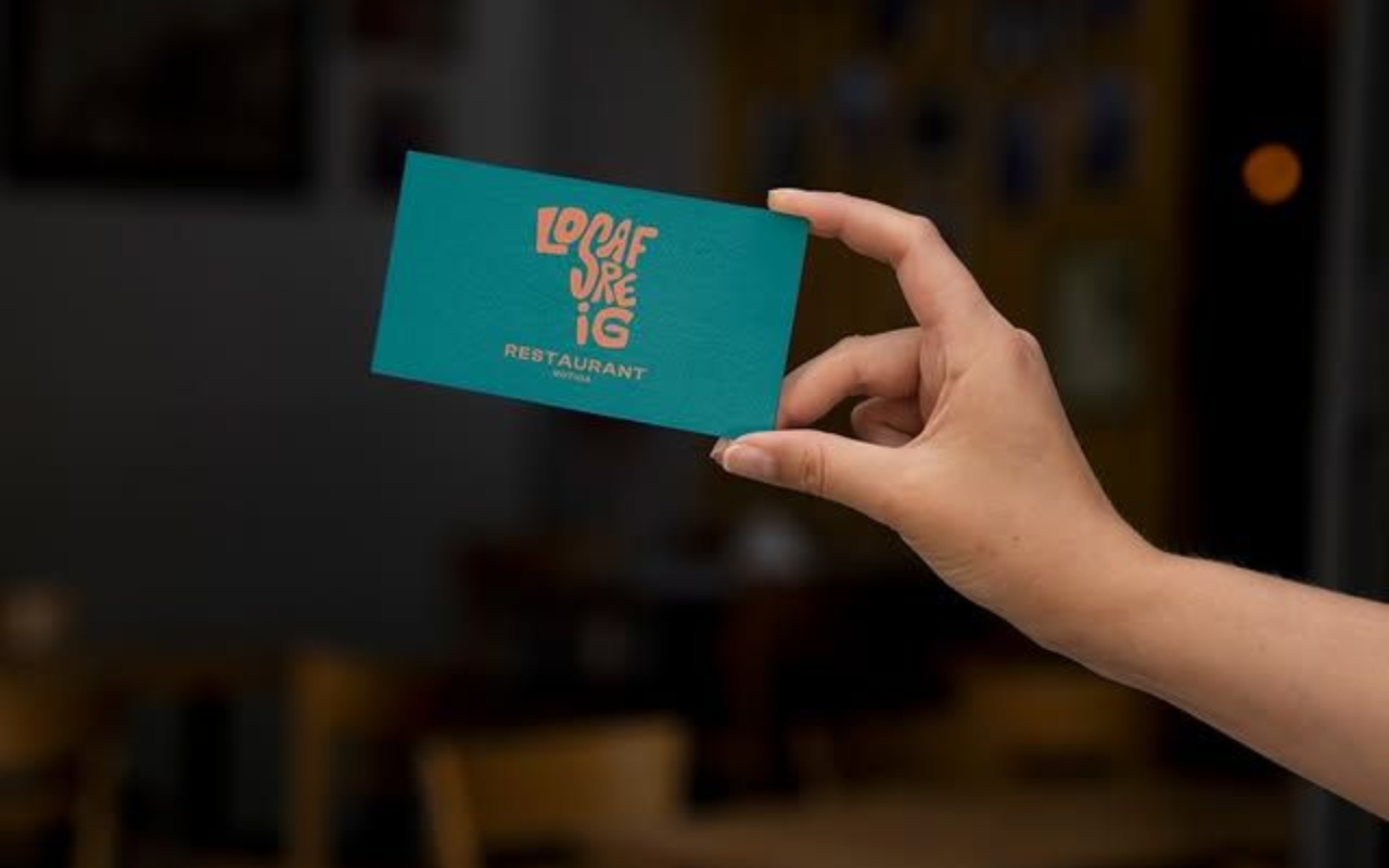 Mano sosteniendo una tarjeta de visita color verde azulado con texto dorado dentro de un restaurante.