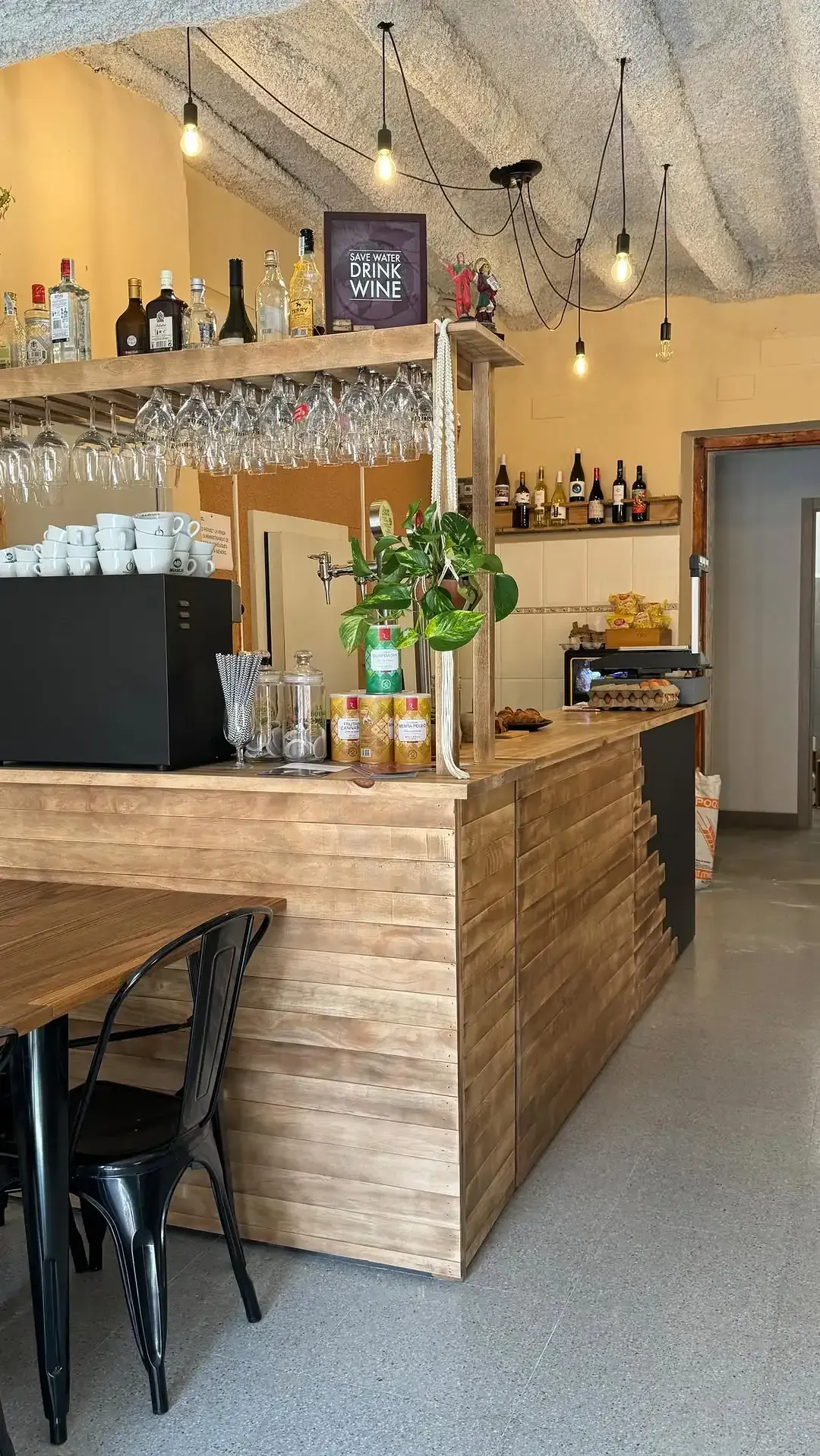 Interior de cafetería con barra de madera, lámparas colgantes y mesa con sillas.