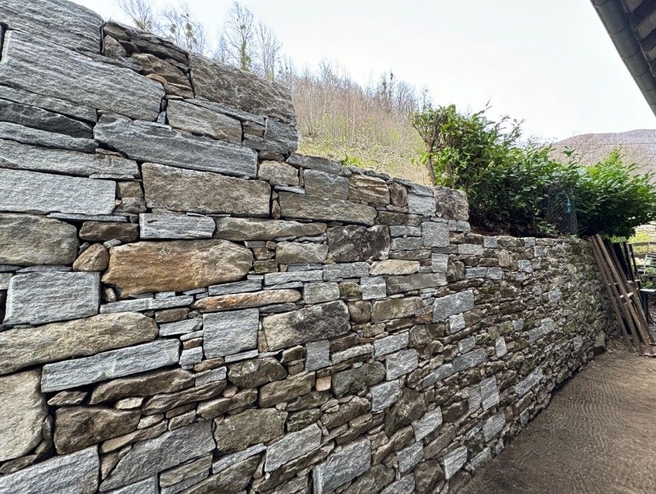 Casa rossa con dettagli in pietra, piccolo ruscello e montagne sullo sfondo. Giornata di sole.