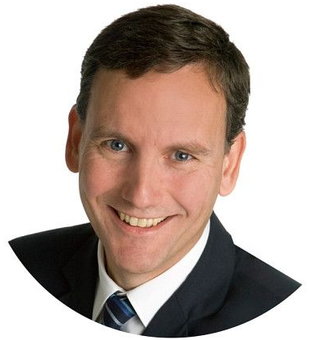 Mann im dunklen Anzug lächelt; helle Haut, blaue Augen, weißer Hintergrund.