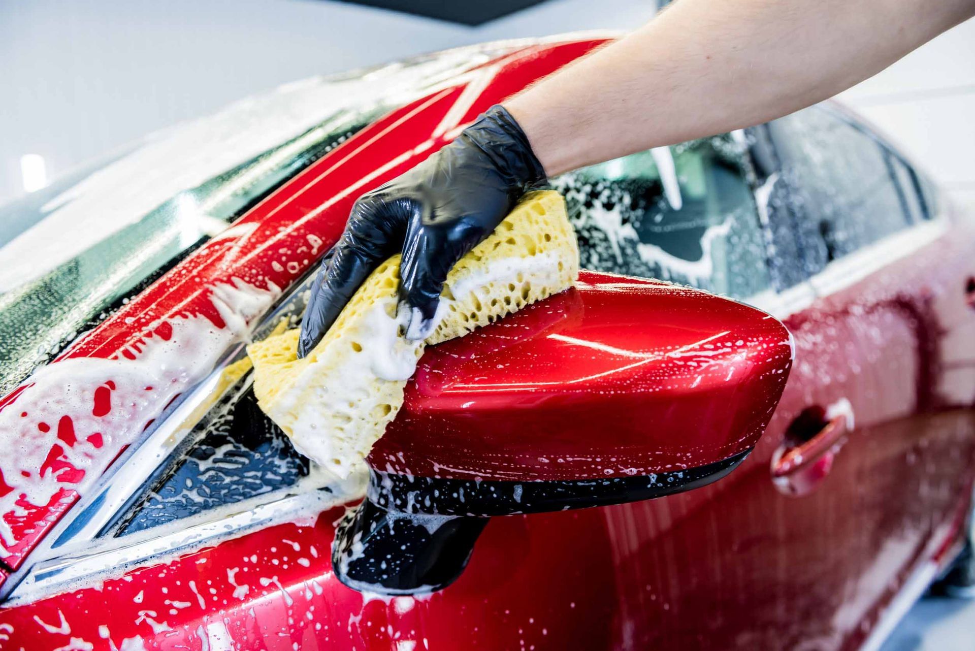 Persona lavando el espejo lateral de un coche rojo con una esponja jabonosa y usando un guante negro.