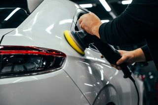 Una persona pule un coche blanco con una pulidora rotativa en un garaje bien iluminado.