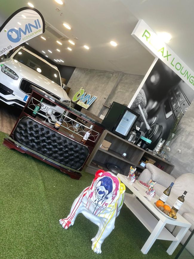 Salón interior con una camioneta blanca, una estatua de bulldog y una mesa con bebidas. Suelo de césped verde e iluminación cenital.