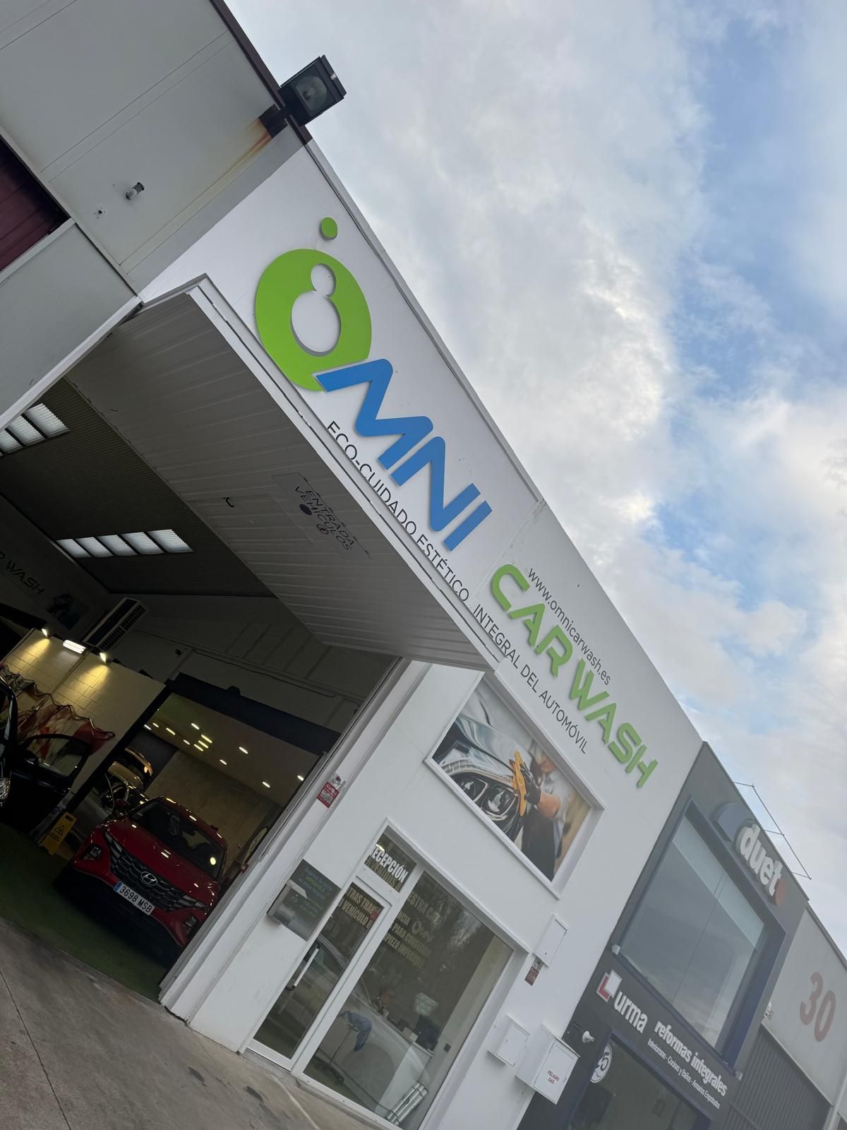Edificio Omni Car Wash de color blanco con un logotipo verde y azul, bajo un cielo nublado.