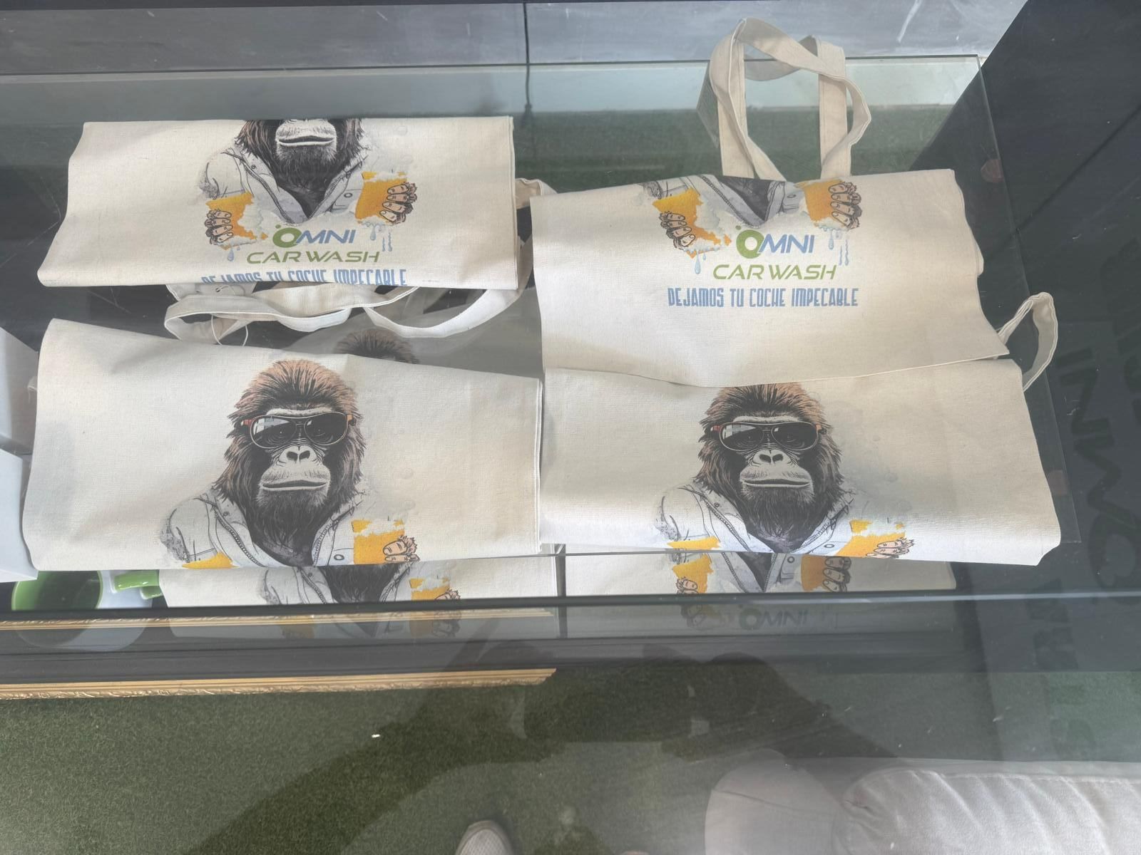 Bolsas de tela y una bolsa doblada con un dibujo de un gorila sosteniendo una cerveza, presentadas en una vitrina. Las bolsas tienen la inscripción