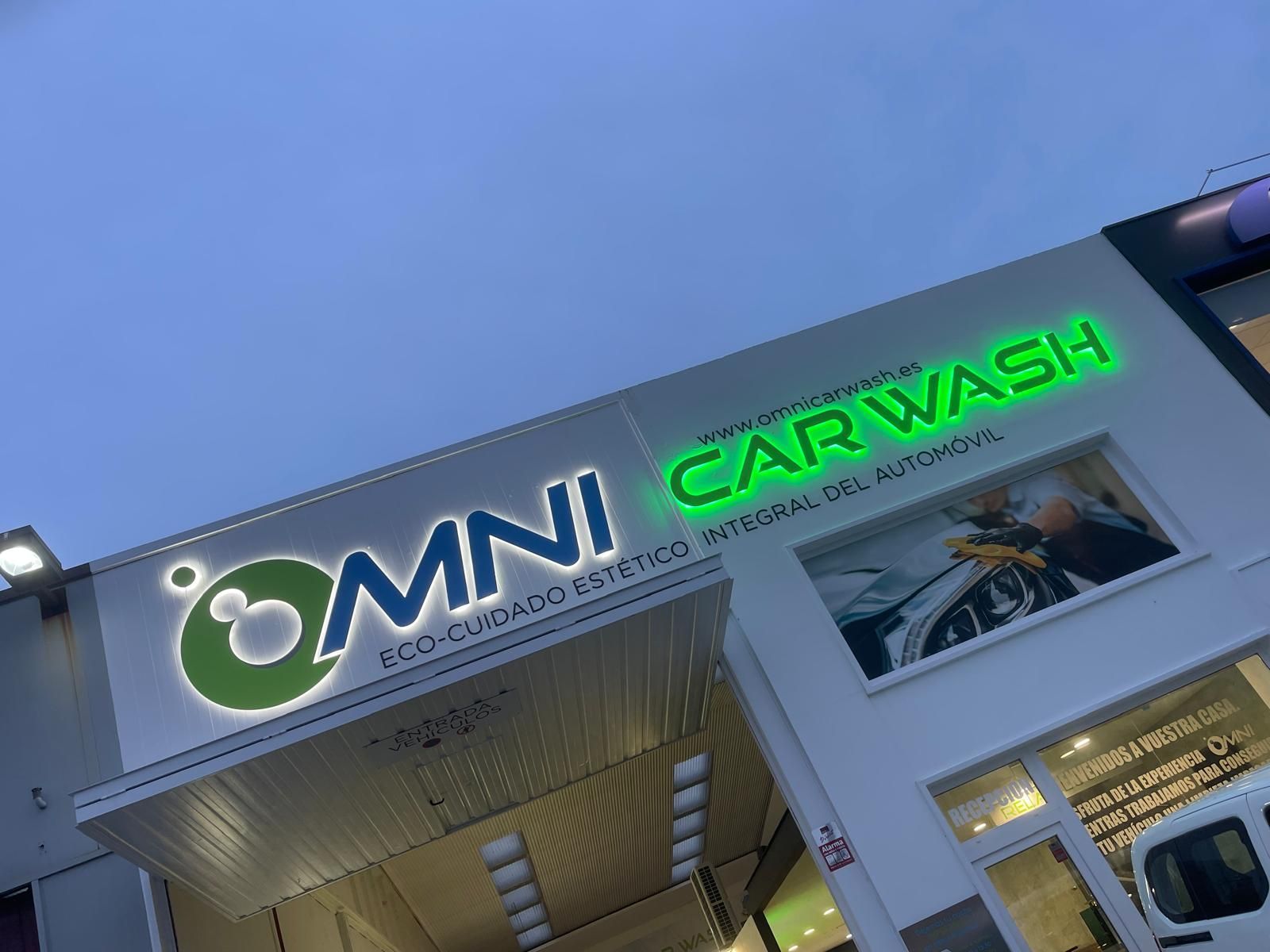 Exterior de “OMNI CAR WASH” con señalización iluminada en verde y un vehículo blanco parcialmente visible.