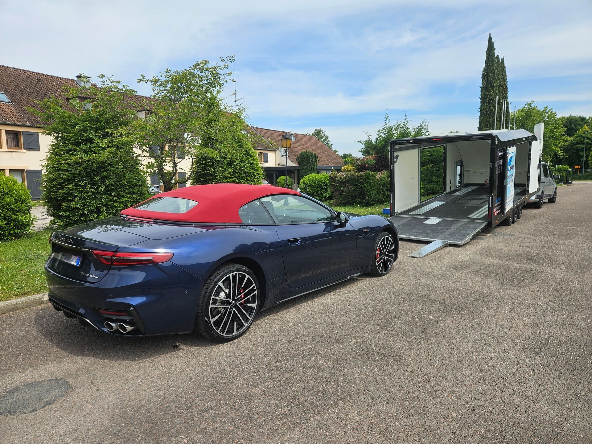 Une voiture de sport décapotable bleu foncé avec un toit rouge est garée sur un parking asphalté devant une remorque.