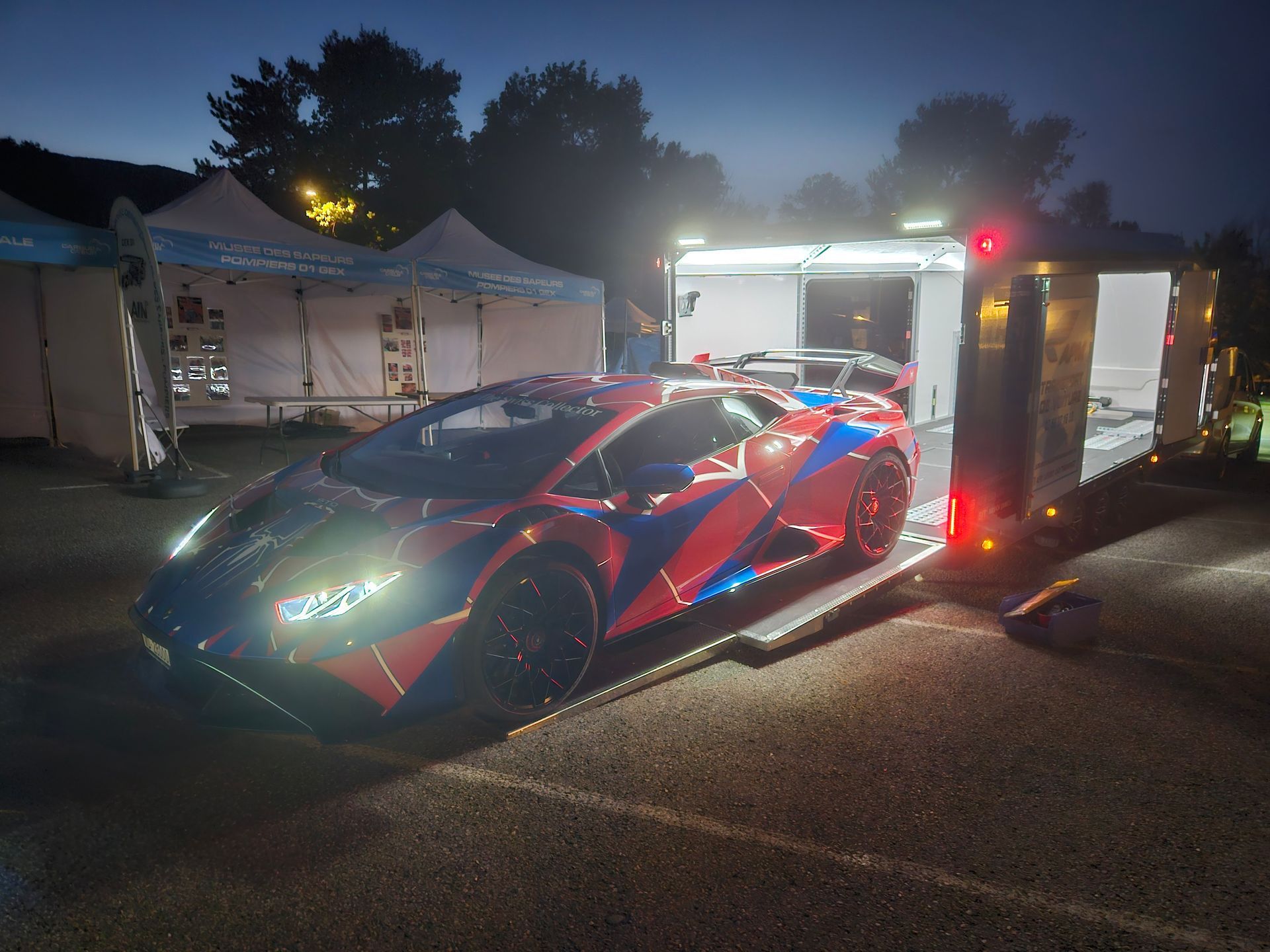 Une Lamborghini à motifs rouges et bleus est chargée sur une remorque, devant des tentes installées pour un événement.