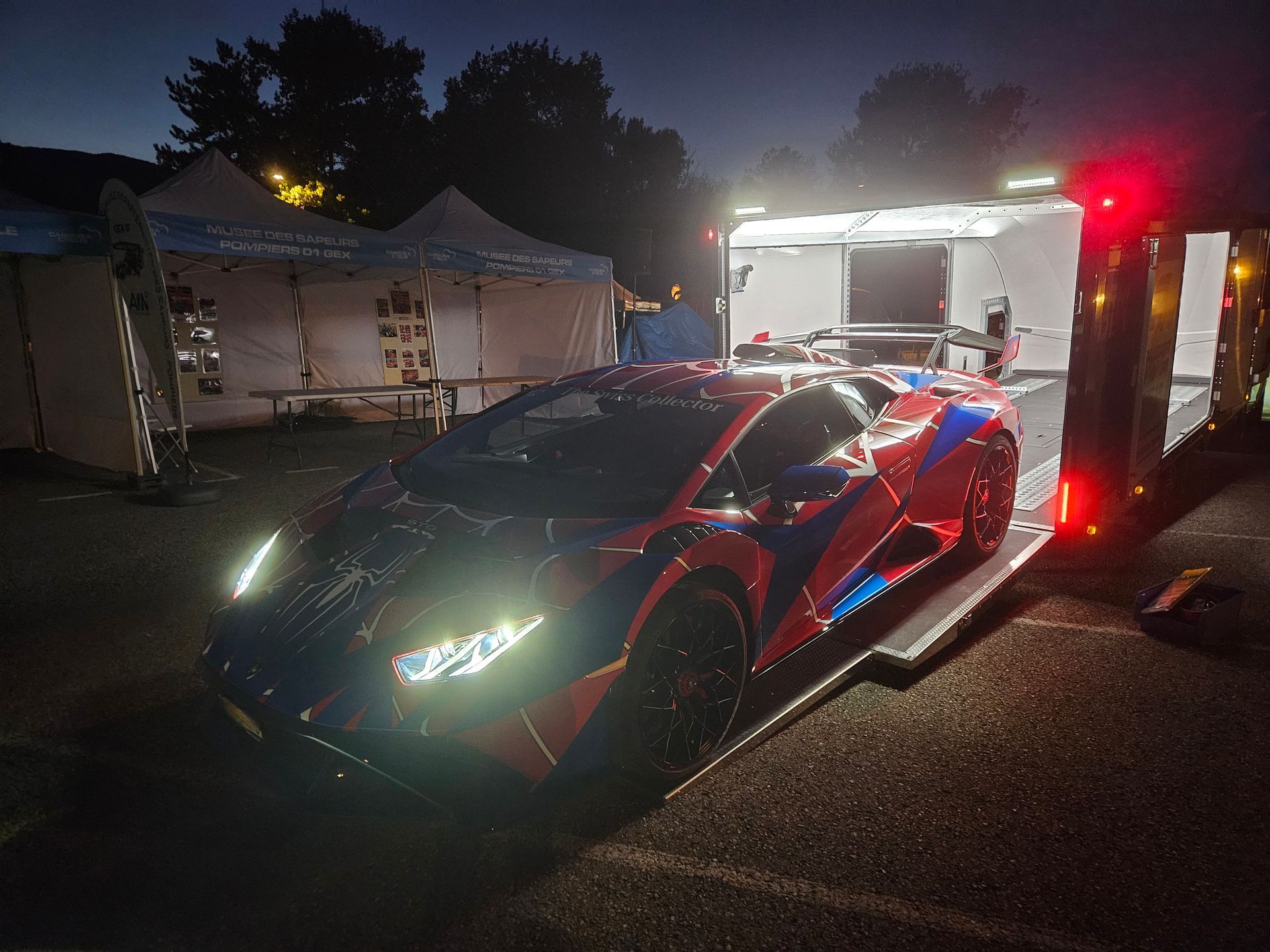 Une Lamborghini à motifs rouges et bleus garée de nuit sur une rampe de remorque, phares allumés.