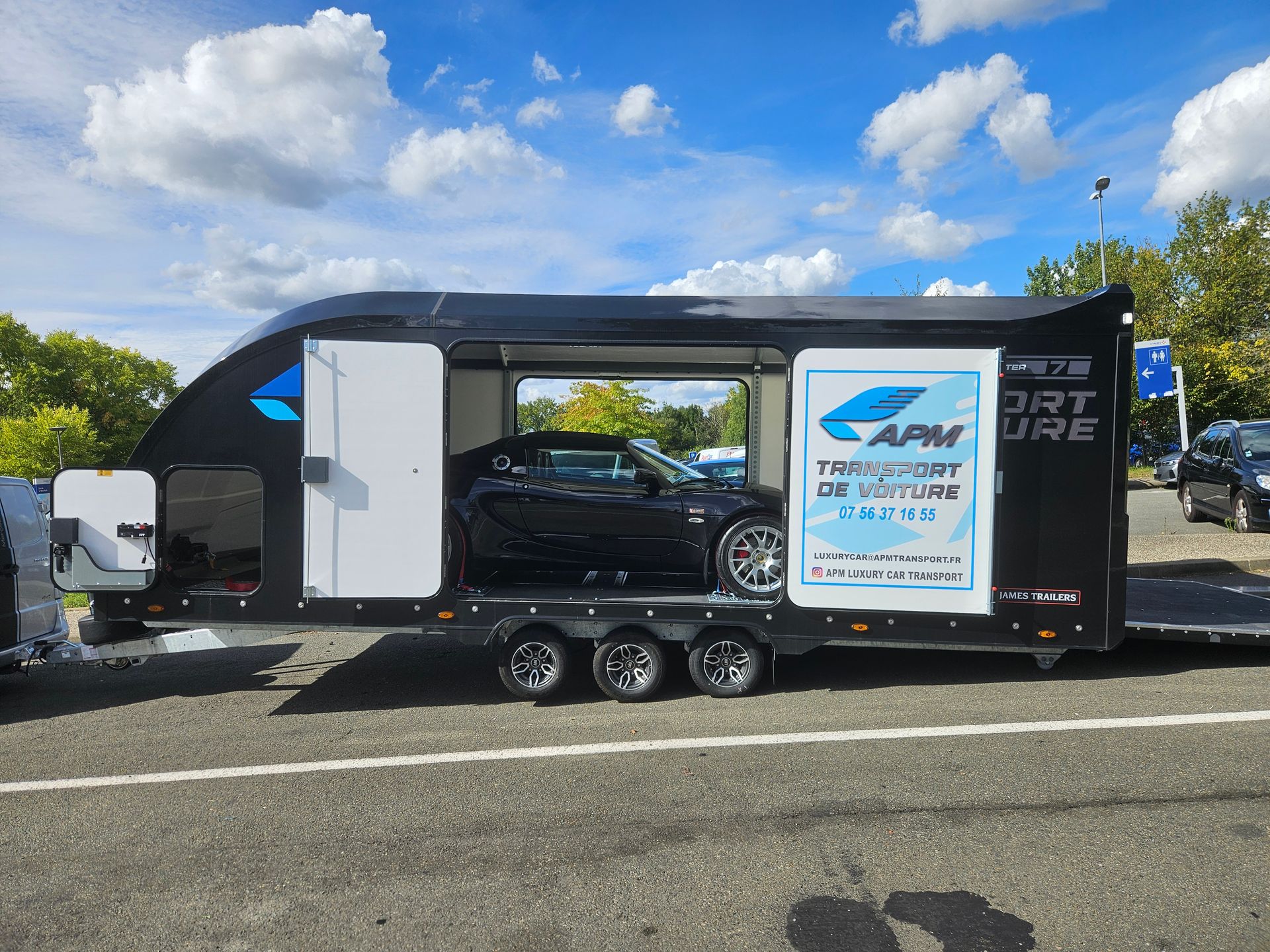 Une remorque porte-voitures noire fermée, avec une porte latérale ouverte, laisse entrevoir une voiture de sport noire garée.