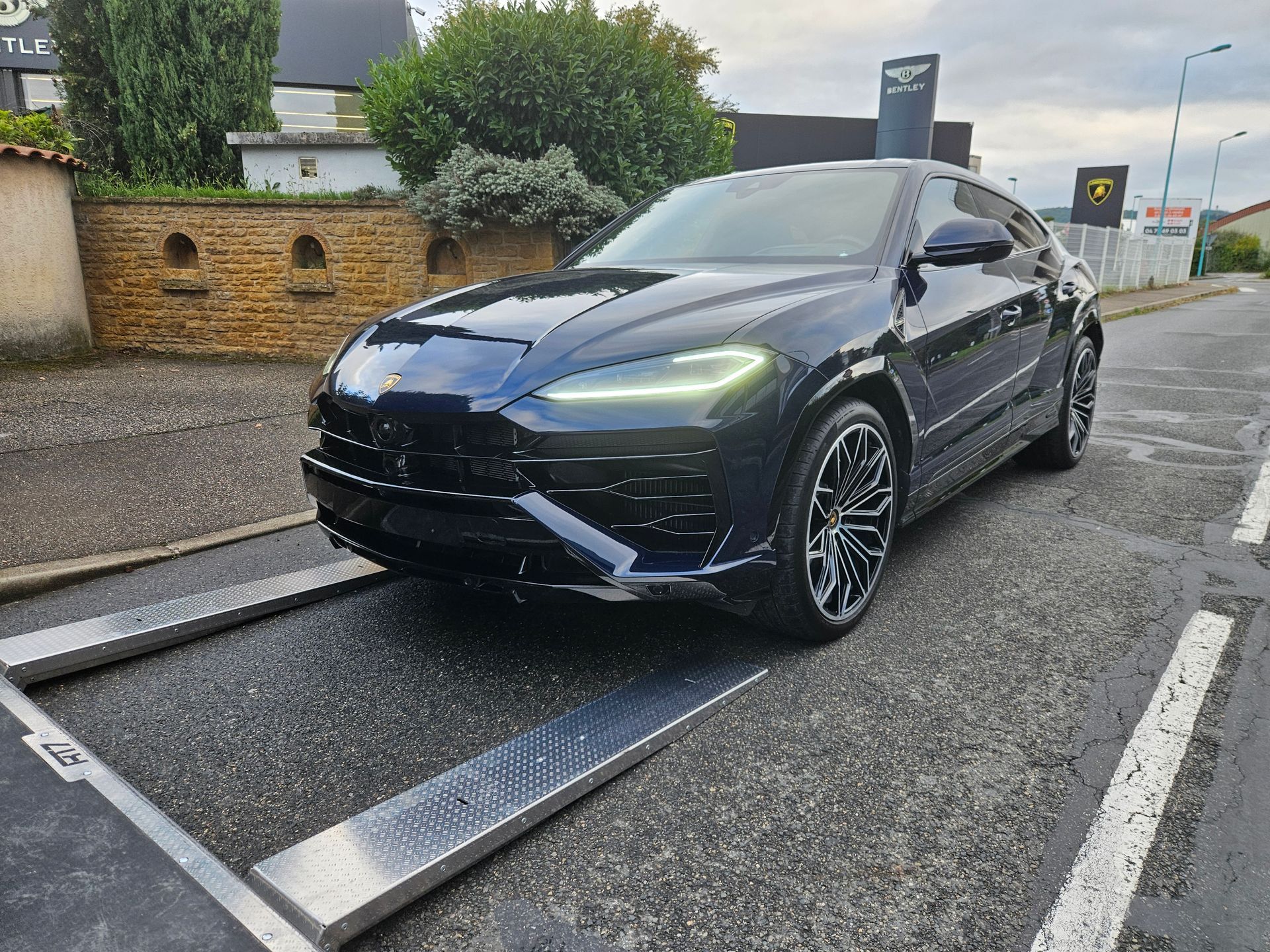 Une Lamborghini Urus de couleur sombre est garée sur une balance métallique à l'extérieur, près d'un mur de briques.