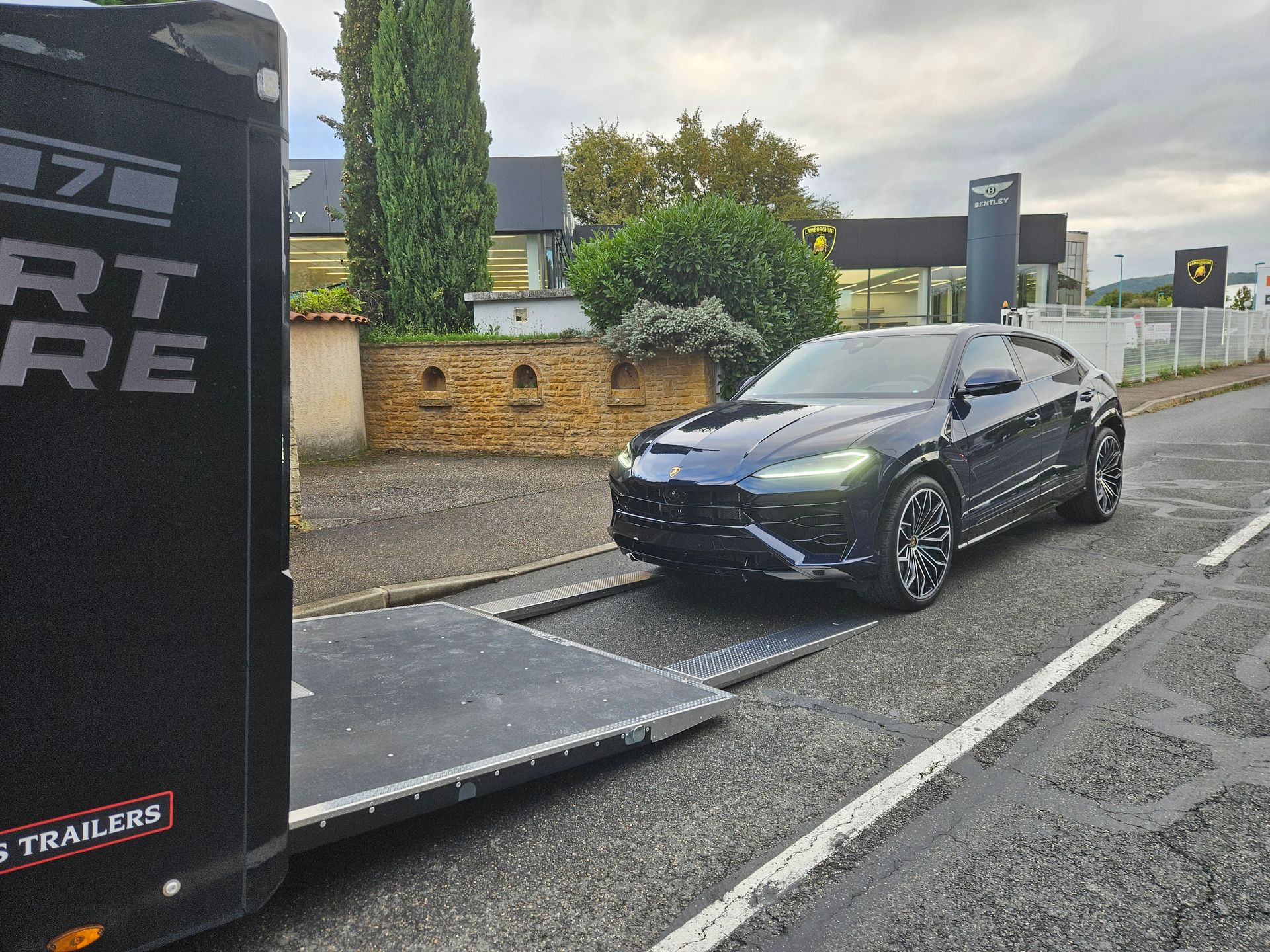 Une Lamborghini Urus bleu foncé est garée sur une remorque porte-voitures devant une concession Lamborghini.