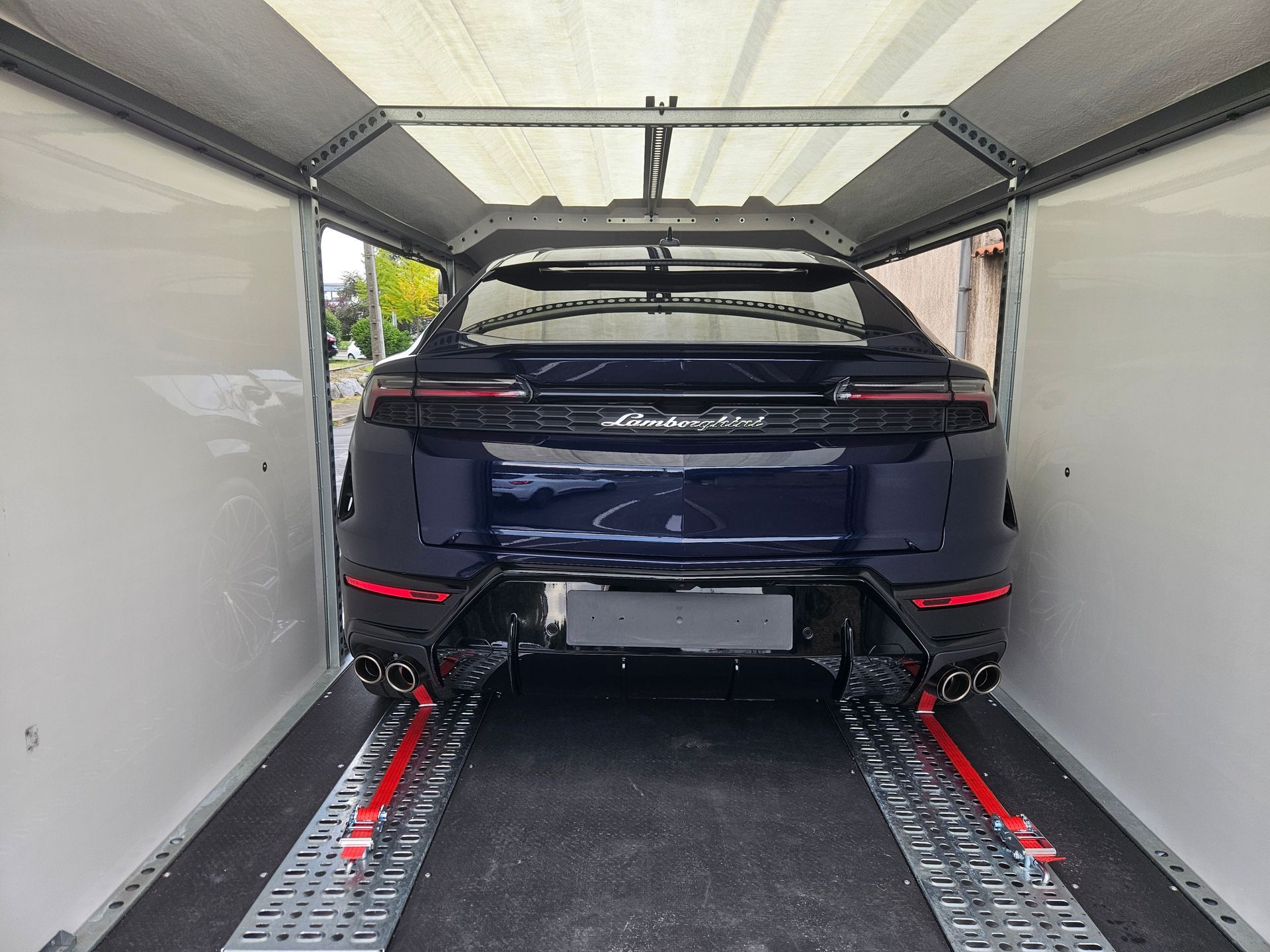 Un SUV Lamborghini Urus bleu foncé, arrimé par des sangles rouges à l'intérieur d'une remorque porte-voitures fermée.
