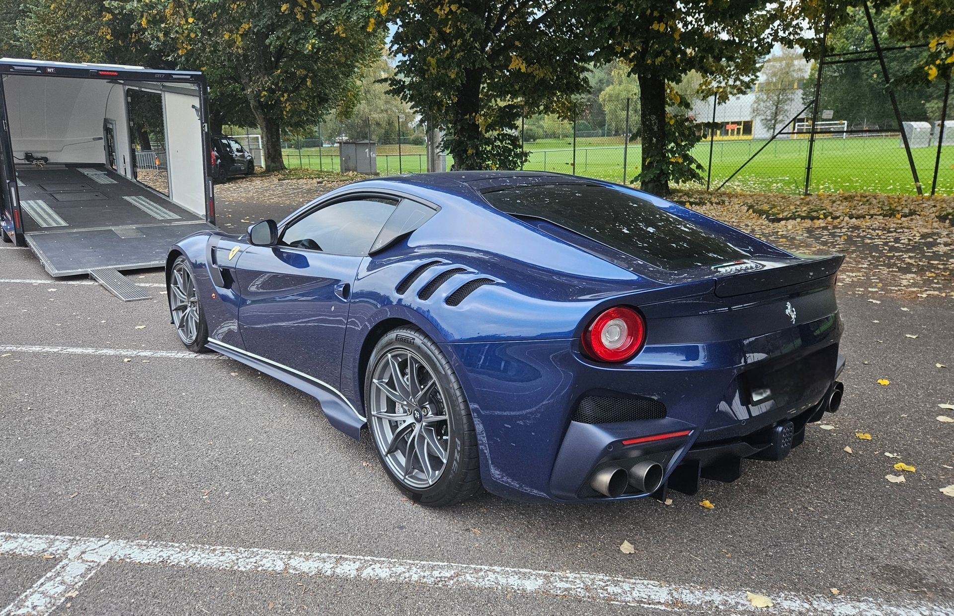 Une Ferrari bleu métallisé garée sur l'asphalte à côté d'une remorque ouverte, sur un terrain bordé d'arbres.