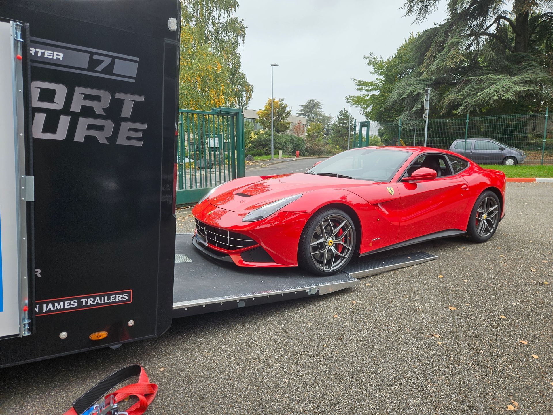 Une Ferrari rouge garée sur une remorque à plateau à côté d'une remorque noire, avec un parking en gravier en arrière-plan.