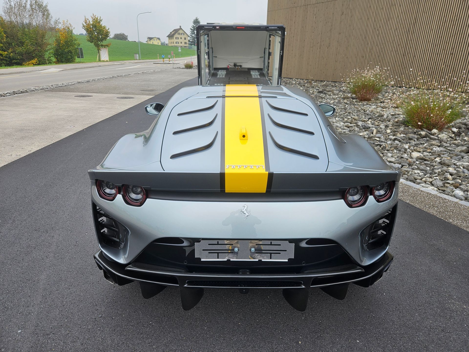 Vue arrière d'une Ferrari 812 Competizione grise avec une bande de course jaune, garée sur une route asphaltée.