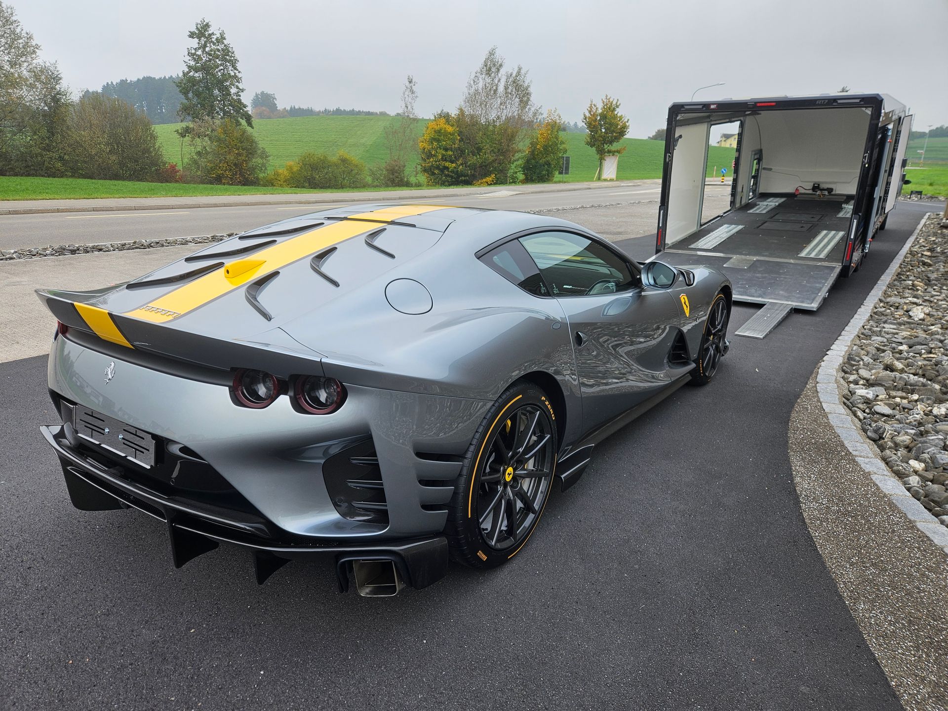 Une Ferrari 812 Competizione grise avec une bande de course jaune est garée à côté d'une remorque porte-voitures ouverte.