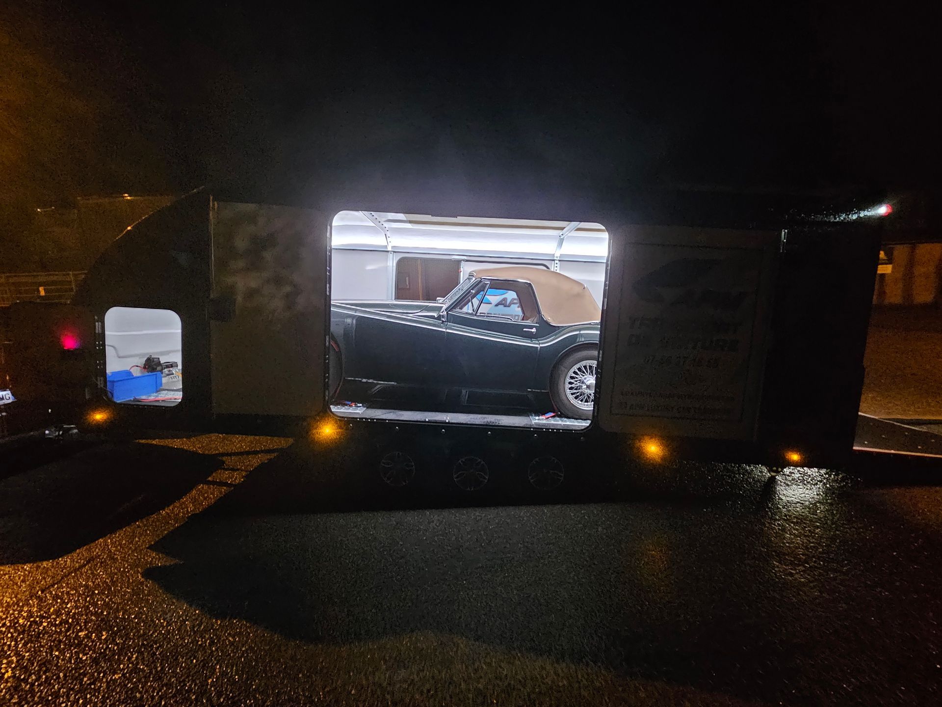 Une voiture décapotable de couleur sombre garée de nuit à l'intérieur d'une remorque porte-voitures ouverte.