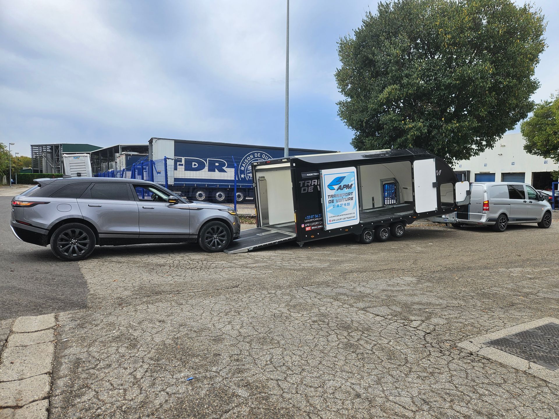 Un Range Rover argenté garé à côté d'une remorque porte-voitures ouverte sur un parking, avec une camionnette blanche.