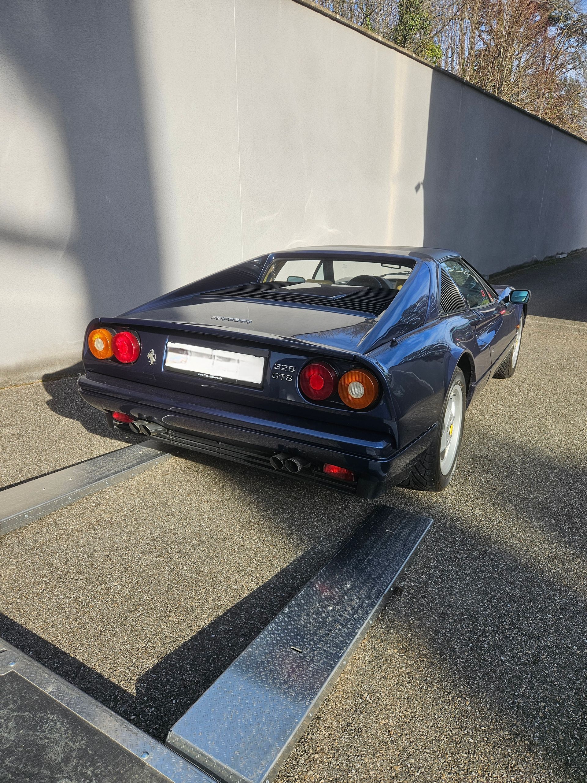 Une Ferrari vintage bleu foncé, garée sur une allée de gravier à côté d'un mur en béton.