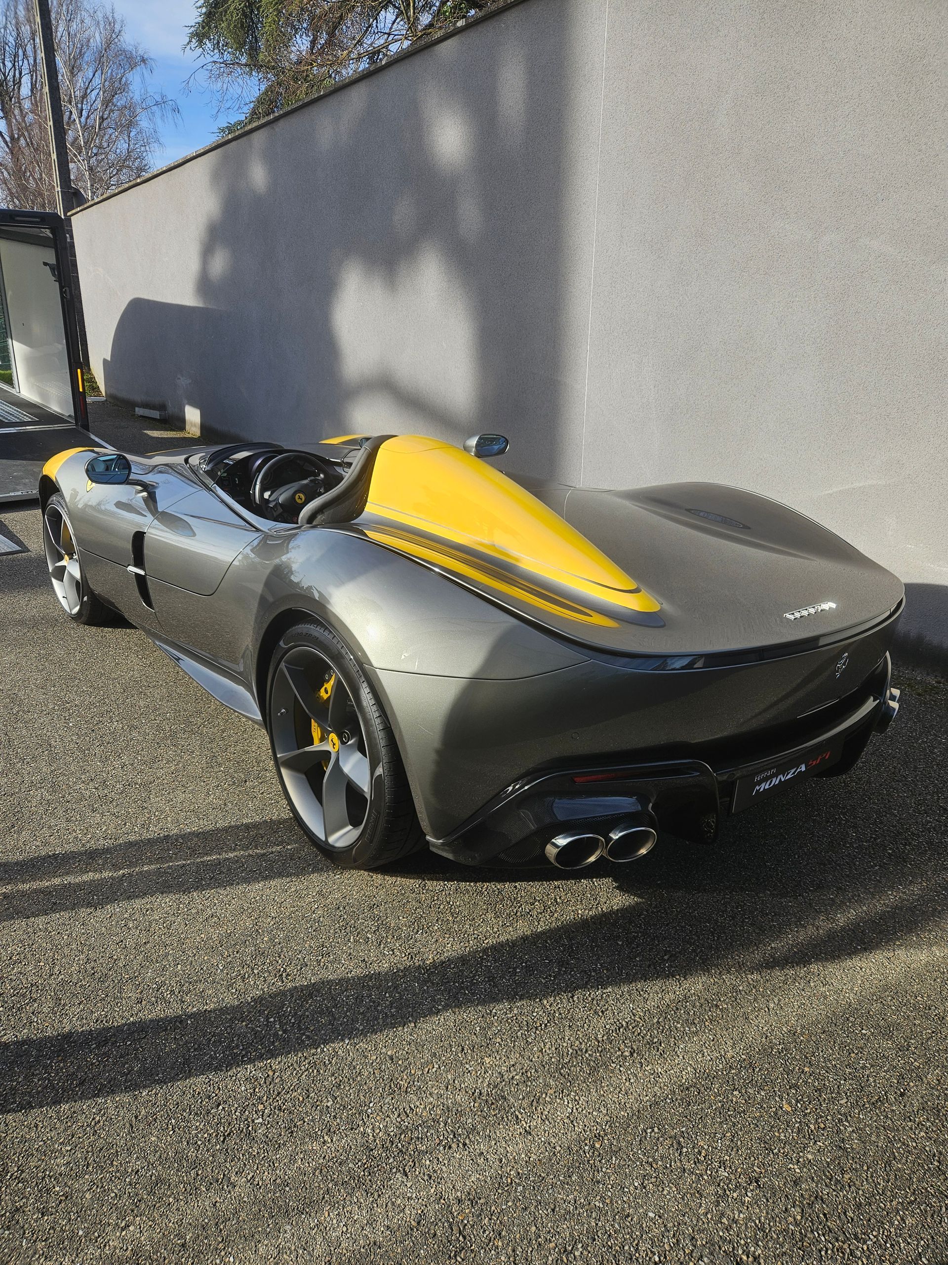 Une supercar Ferrari Monza SP1 grise avec un carénage aérodynamique jaune, garée à l'extérieur sur une surface gravillonnée.