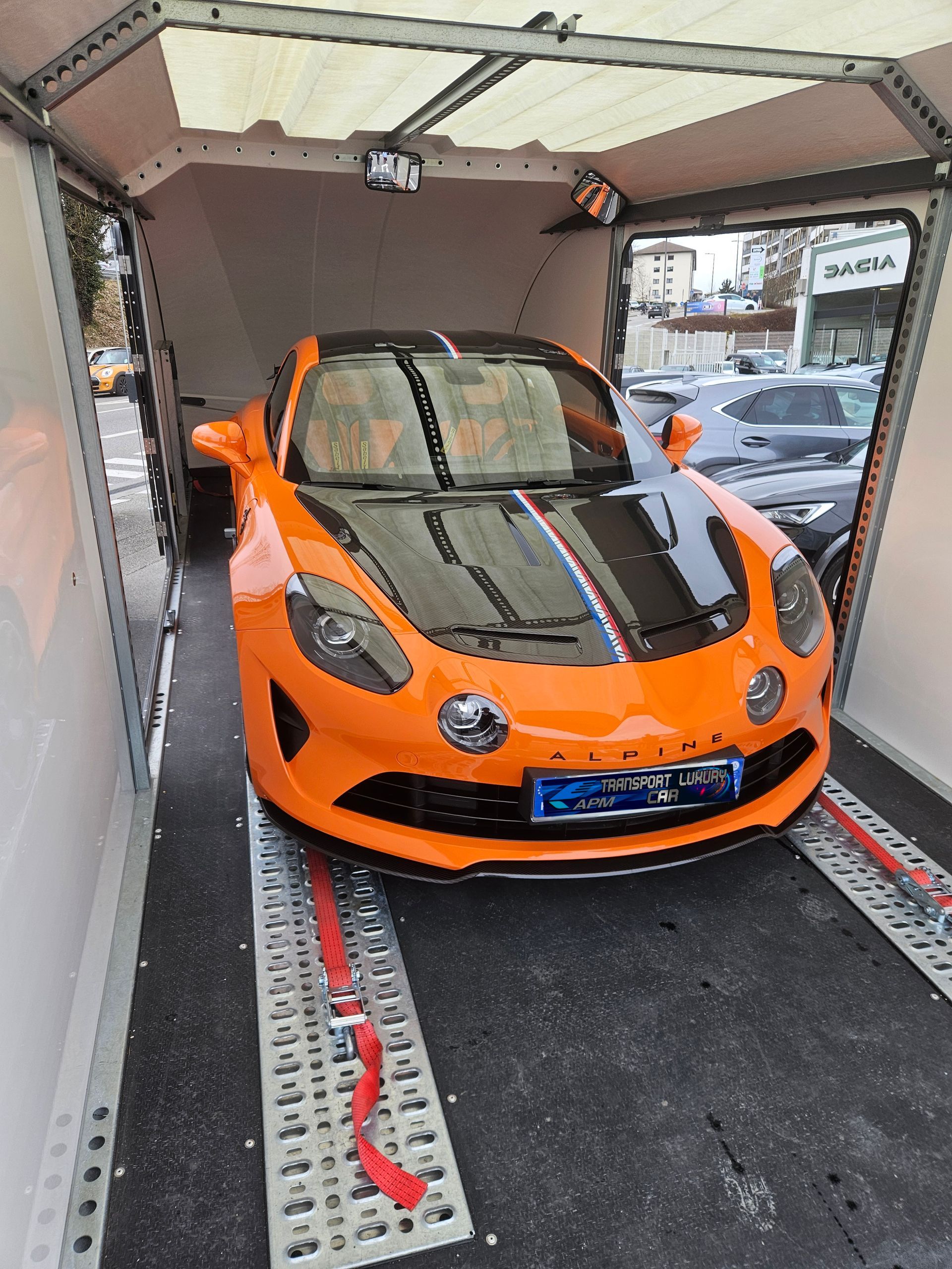 Une voiture de sport Alpine A110 orange, fixée à l'intérieur d'une remorque de transport, avec une bande noire sur le capot.