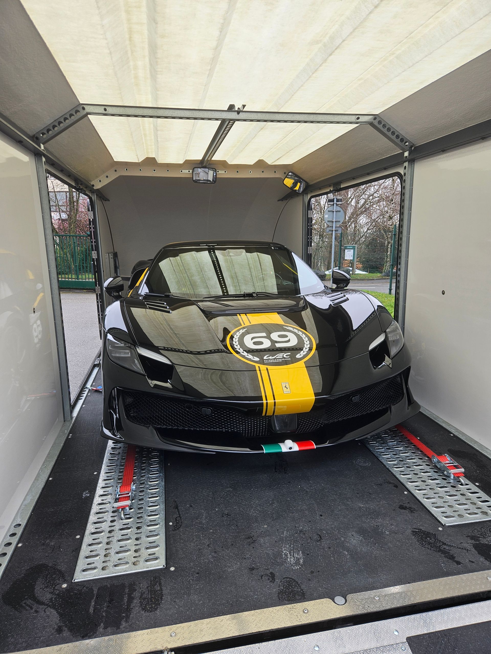 Une Ferrari noire avec une bande de course jaune et le numéro 69 dans une remorque porte-voitures fermée.