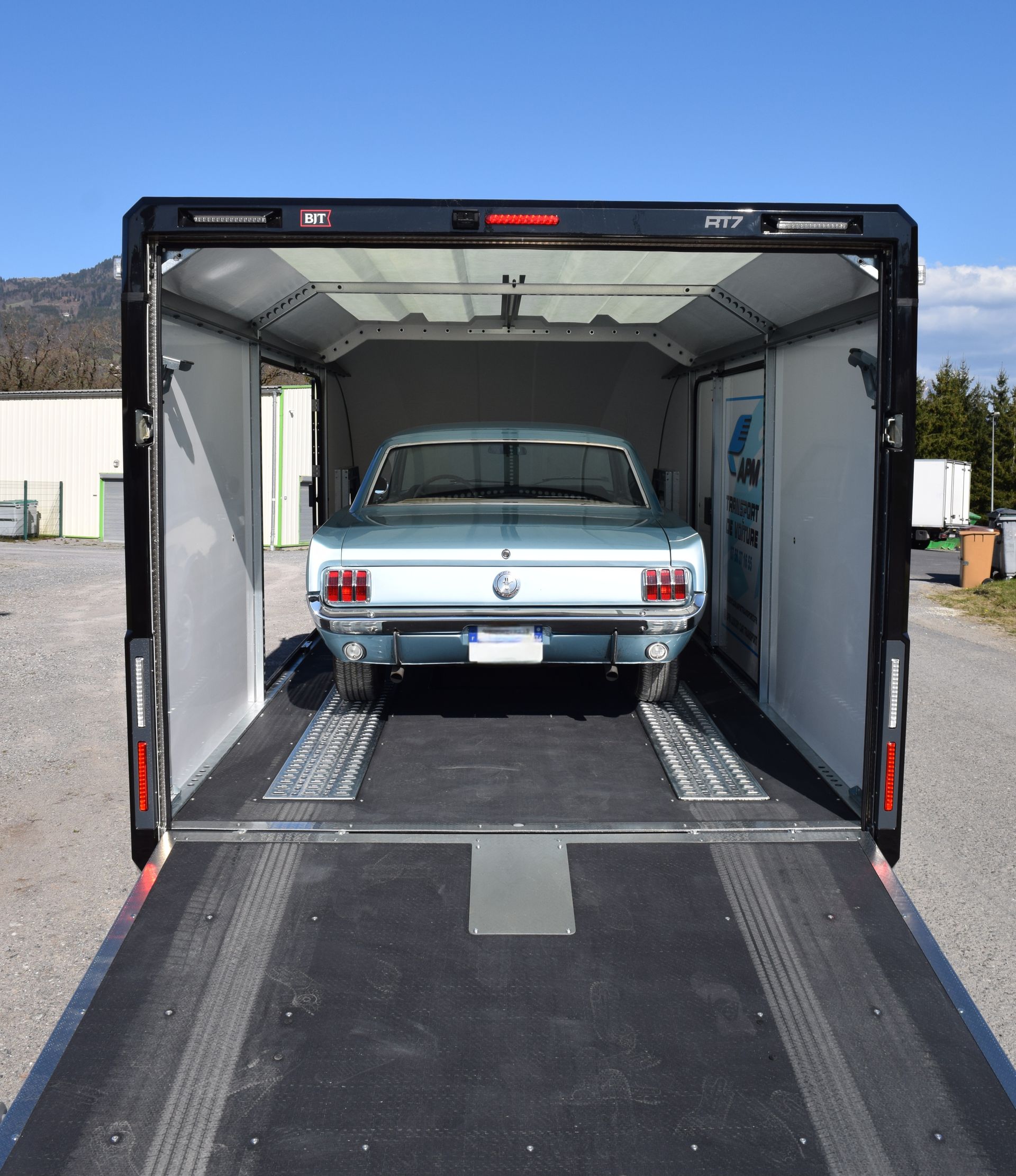 Une voiture ancienne bleu clair garée à l'intérieur d'une remorque noire fermée, la porte arrière abaissée.