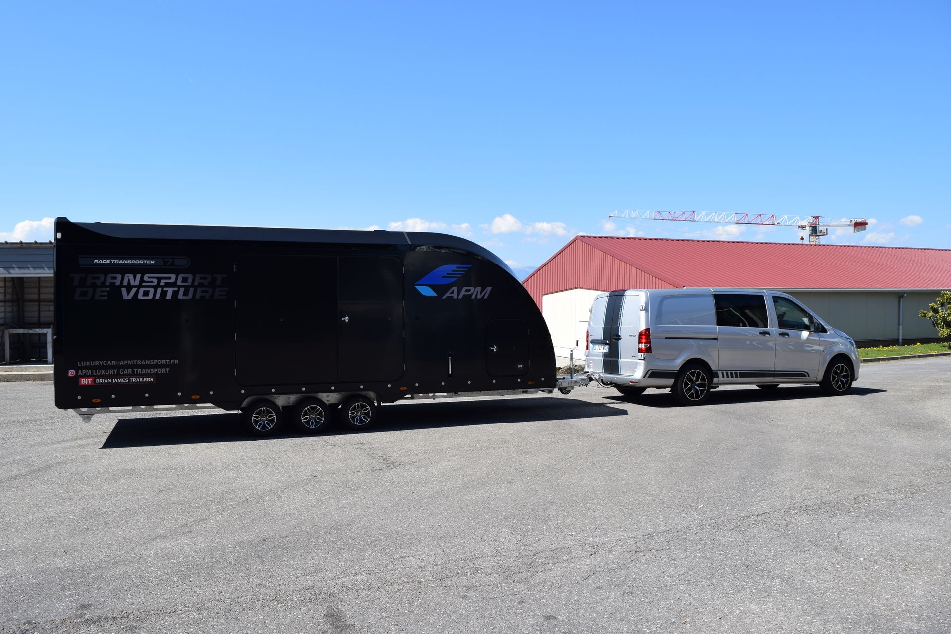Une camionnette tractant une remorque noire fermée est stationnée sur un terrain devant un bâtiment au toit rouge.