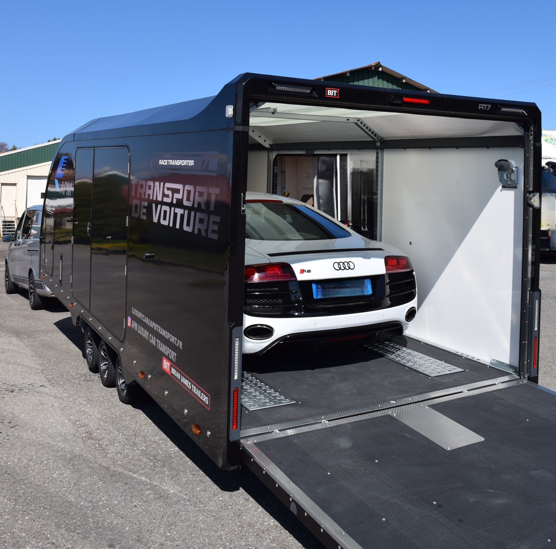 Une Audi R8 argentée est chargée dans une remorque porte-voitures noire et fermée sur un parking asphalté.