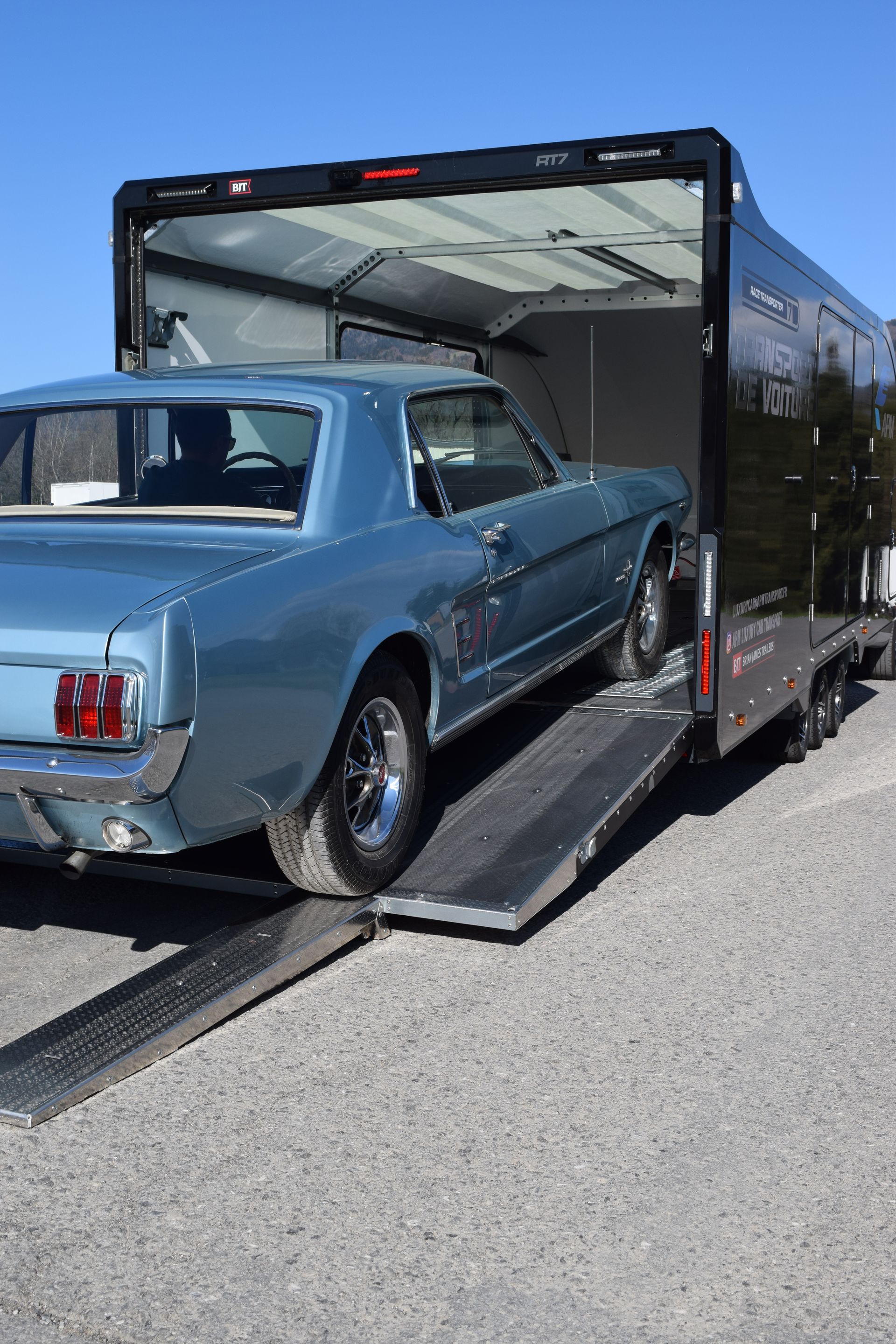 Une Ford Mustang vintage bleu clair est chargée sur une remorque fermée noire à l'aide d'une rampe.